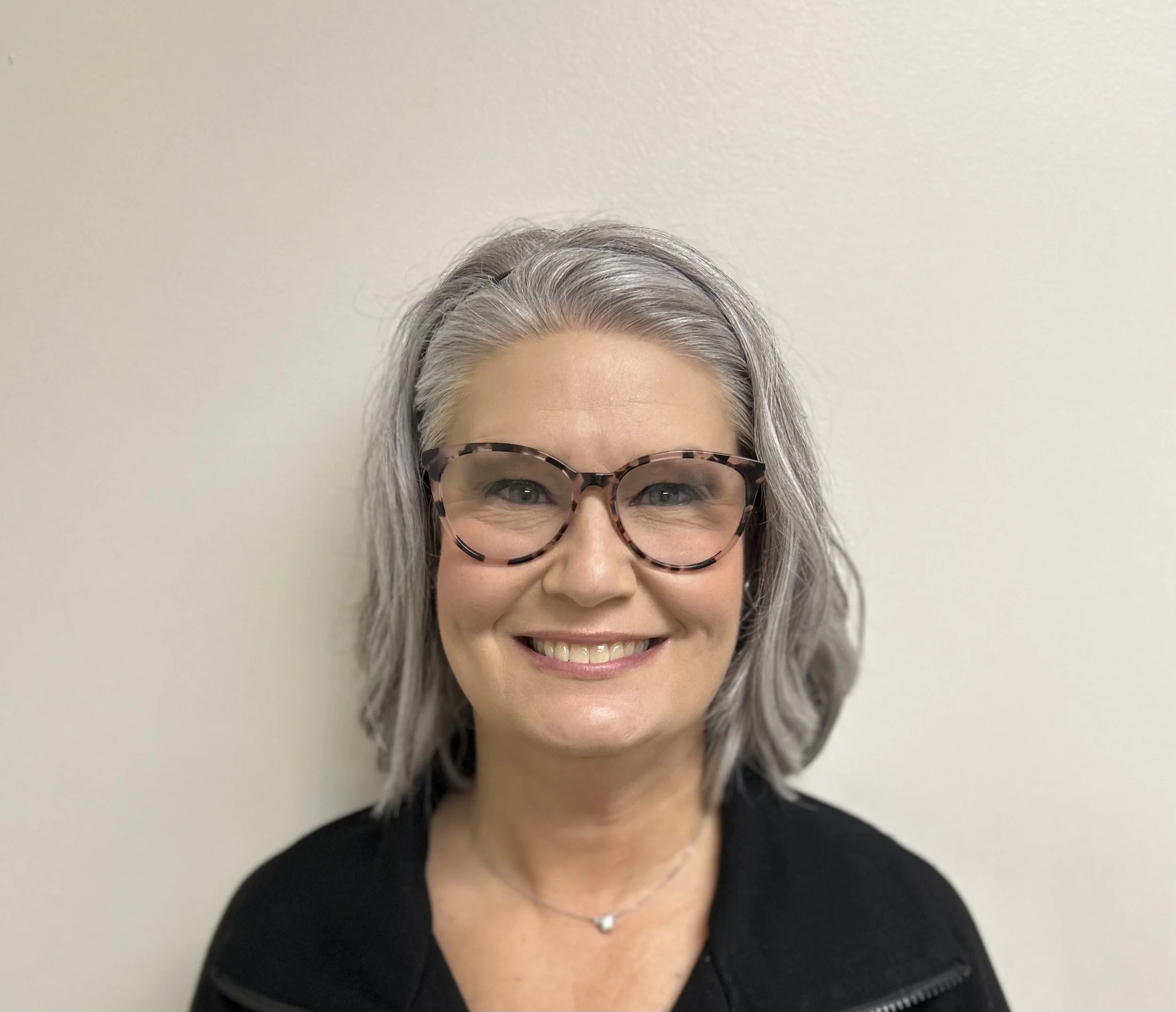 A woman with short gray hair, wearing black glasses, smiling, standing in front of a blue textured wall.