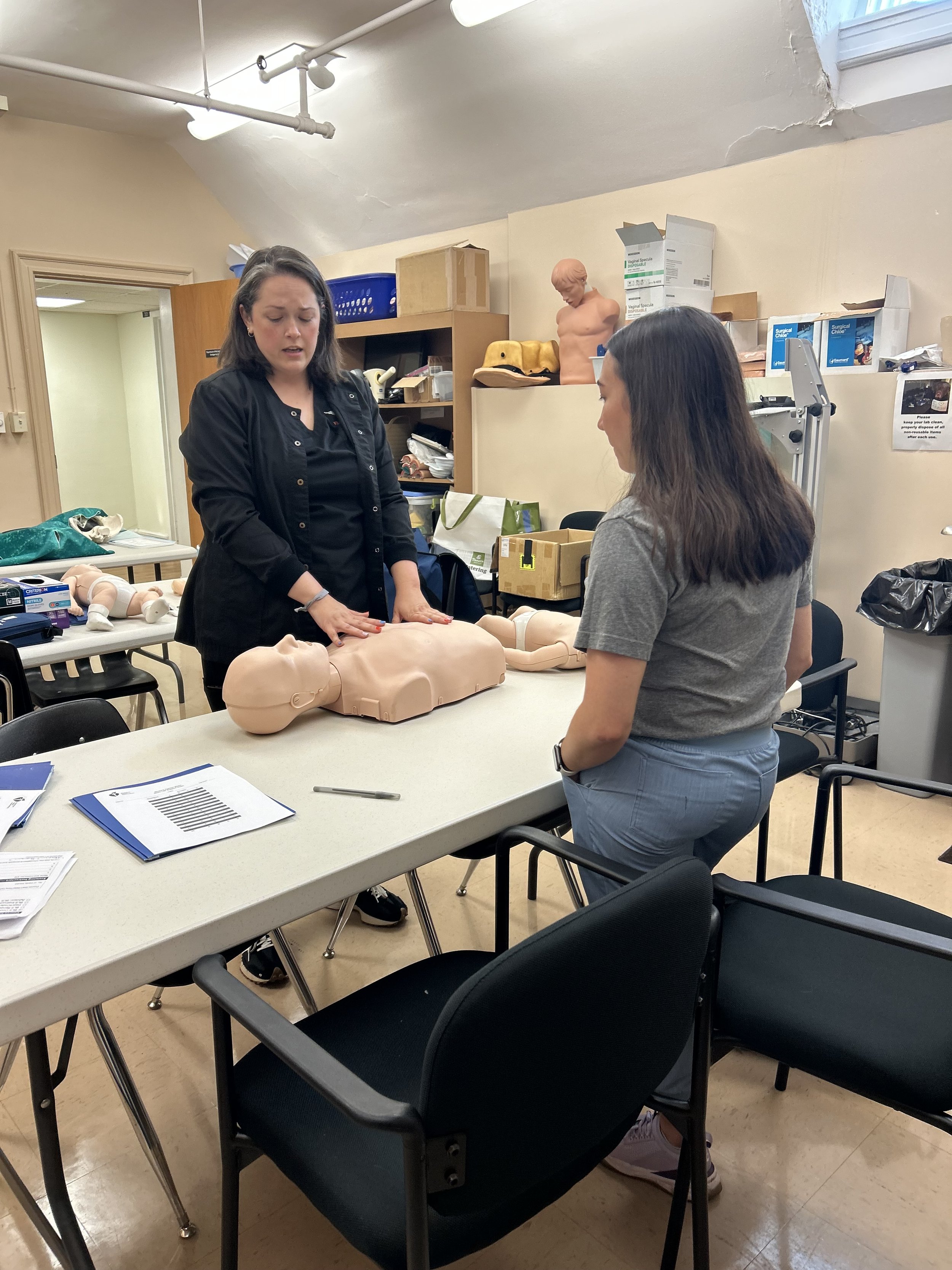 Two women teaching a CPR class using a training mannequin in a classroom. One woman is demonstrating chest compressions, and the other woman is observing.