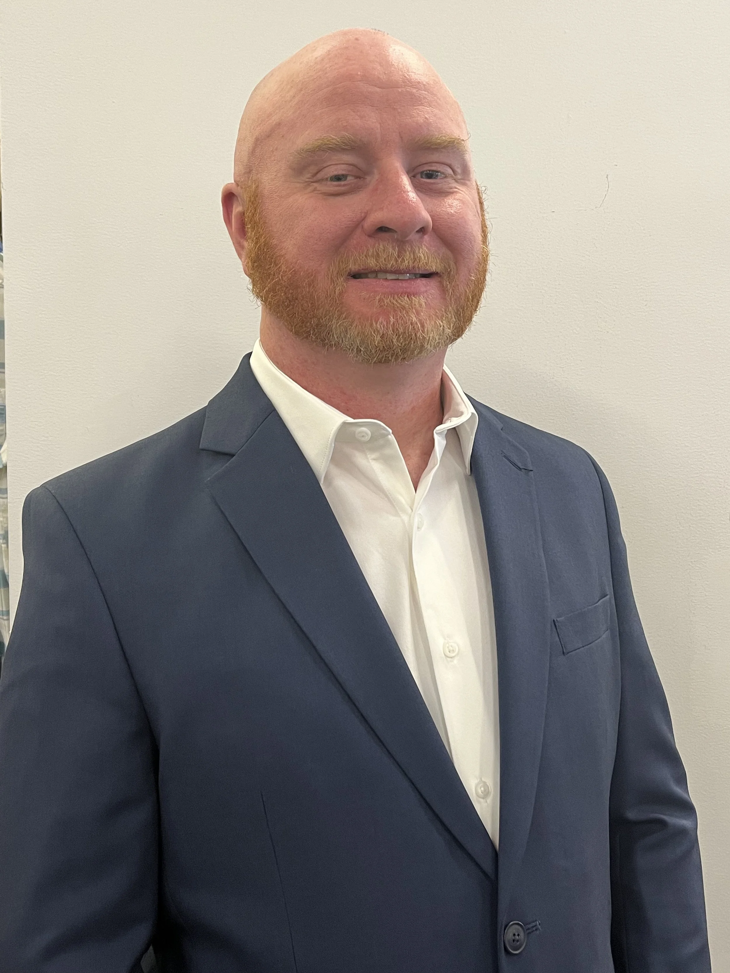 A man with a red beard and bald head wearing a navy blue suit and white shirt standing against a plain beige wall.