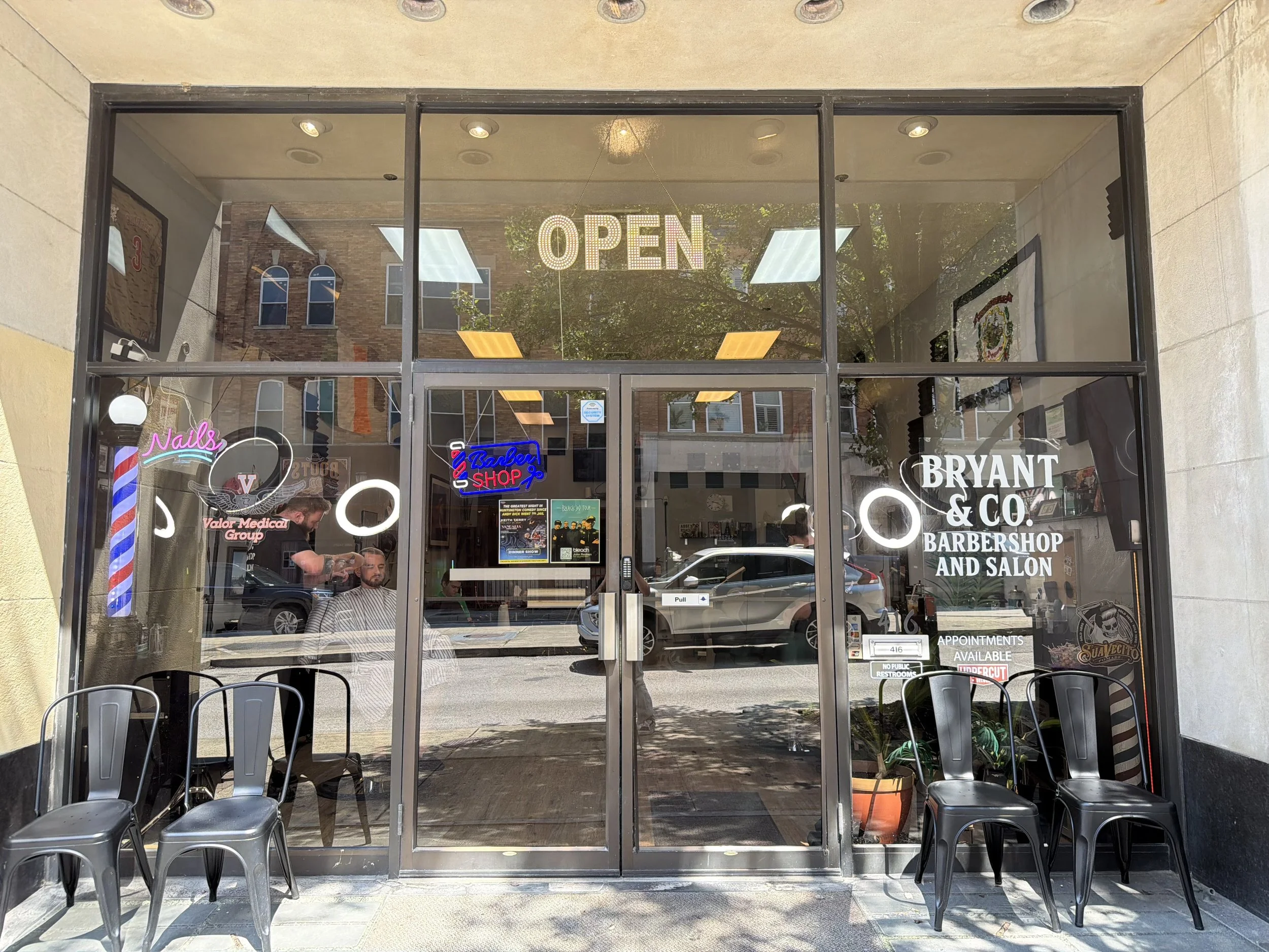 The storefront of Bryant & Co. Barbershop and Salon. Large glass windows and doors reveal interior seating with chairs and customers. An illuminated 'OPEN' sign hangs inside. Various smaller signs and posters are visible on the window, including a neon-style barber shop sign and advertisements for appointments and services.