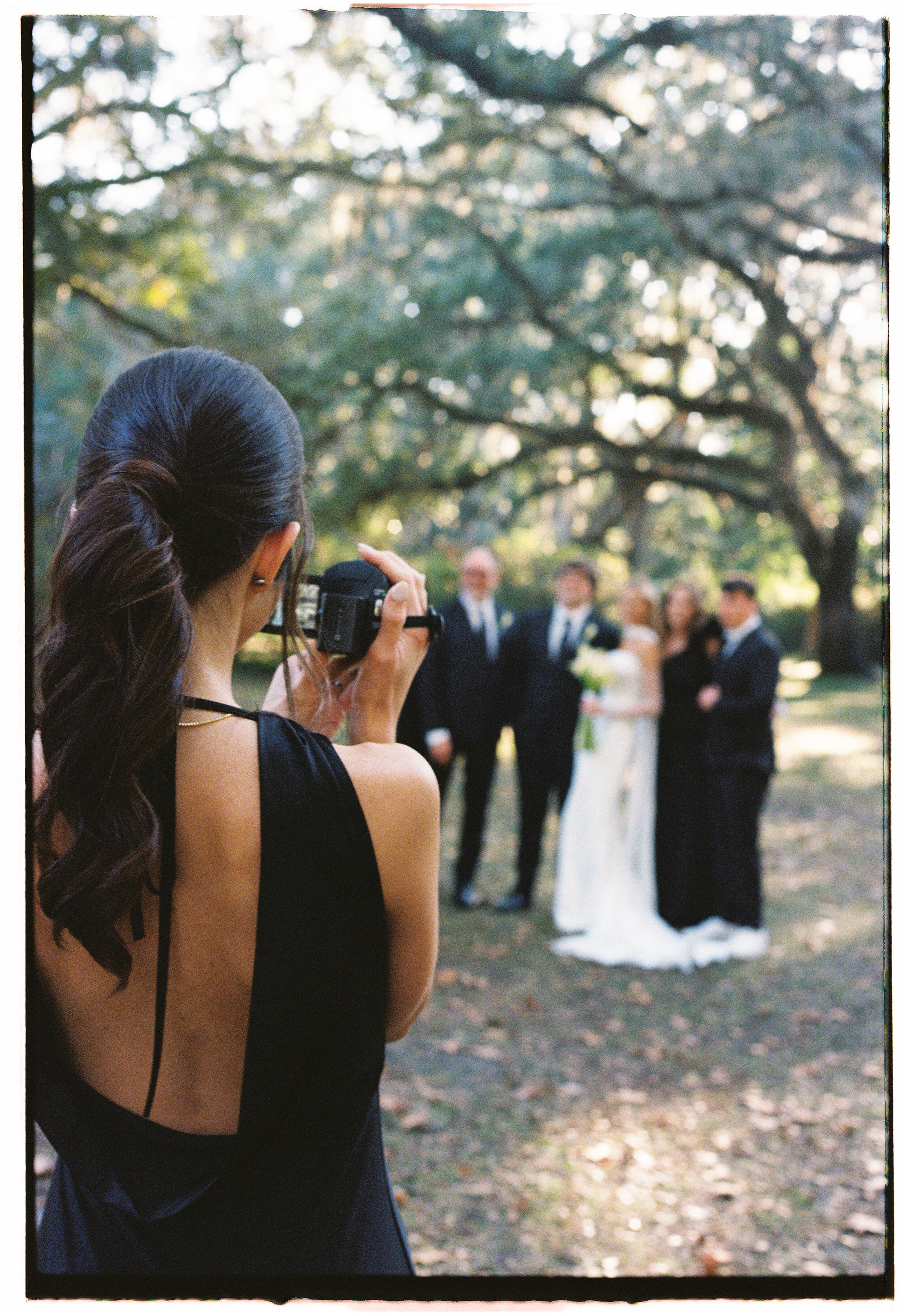 A woman with a dark styled ponytail wearing a sleeveless black dress is taking a photograph of a wedding couple and their guests outdoors under large trees.