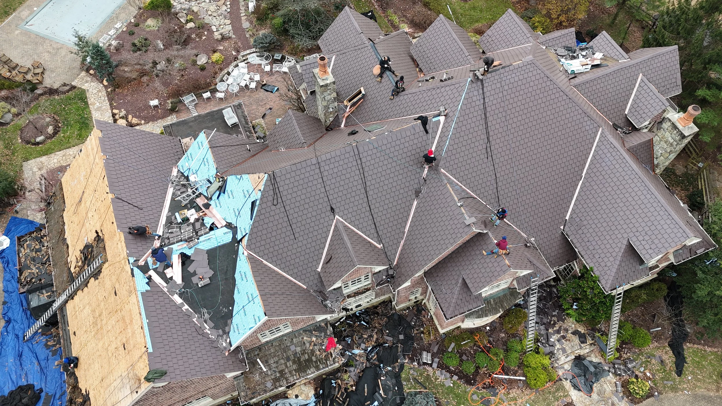 Aerial view of a large house with workers replacing the roof, with some parts under construction and others completed. Roofing materials and ladders are visible.