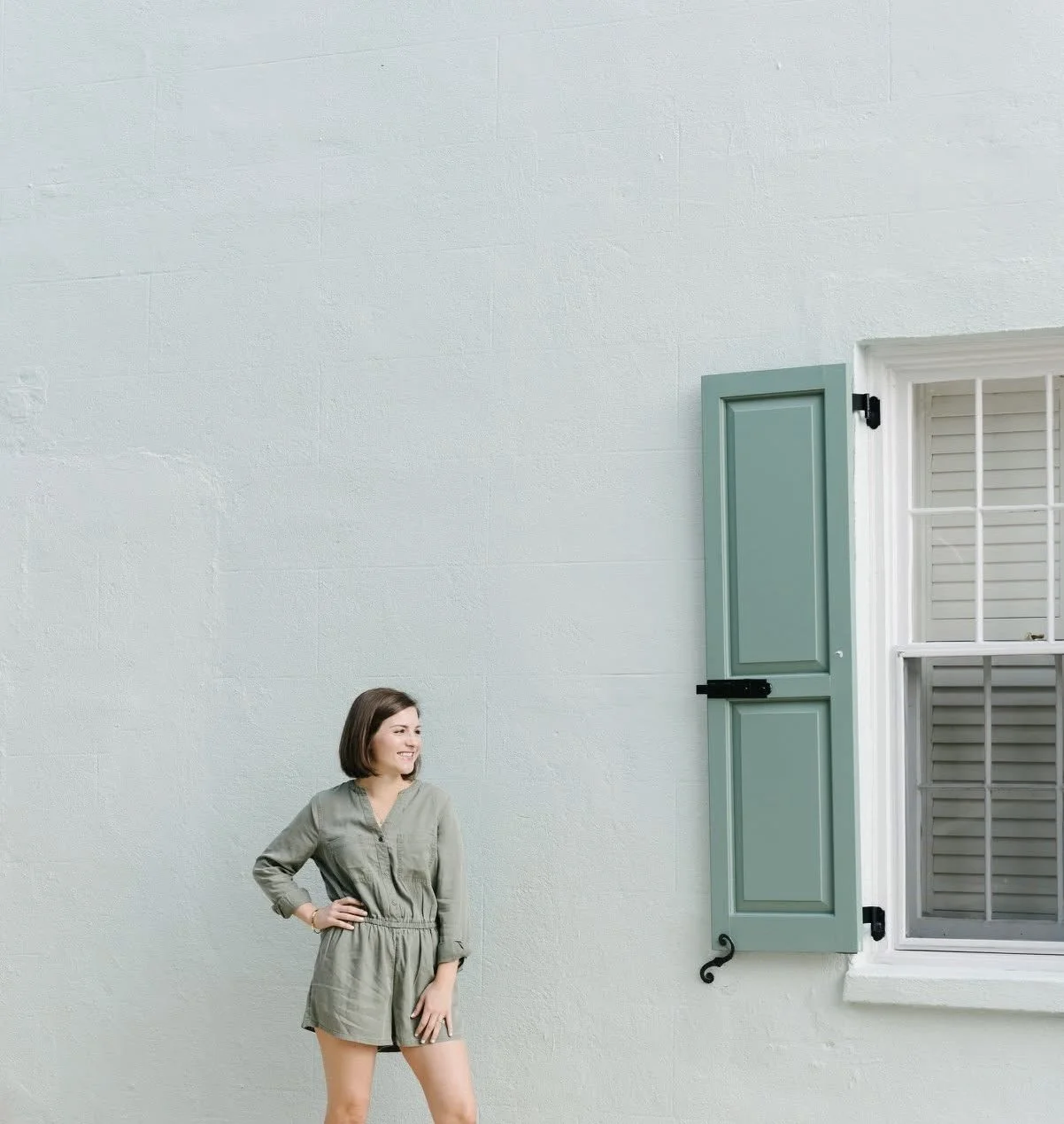 Woman standing outside near a wall with closed window shutters, smiling and looking to her left.