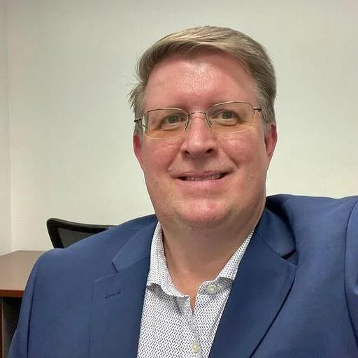 A man with glasses smiling, wearing a blue suit and a white shirt, in an office setting.