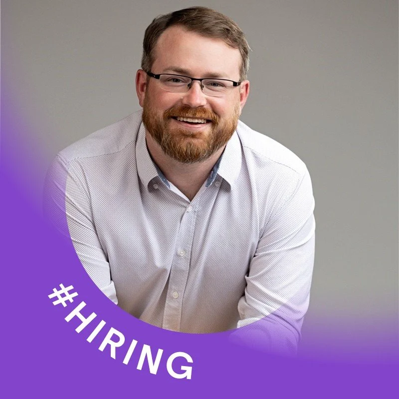 A man with glasses and a beard smiling, wearing a white dress shirt, with a purple circular banner in the lower left corner that says #HIRING.
