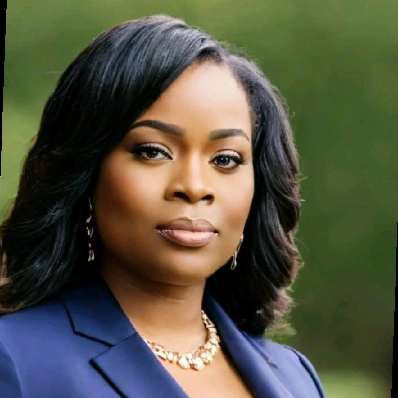 Portrait of an African American woman with wavy black hair, wearing a navy blazer, gold necklace, and earrings, outdoors with a blurred green background.
