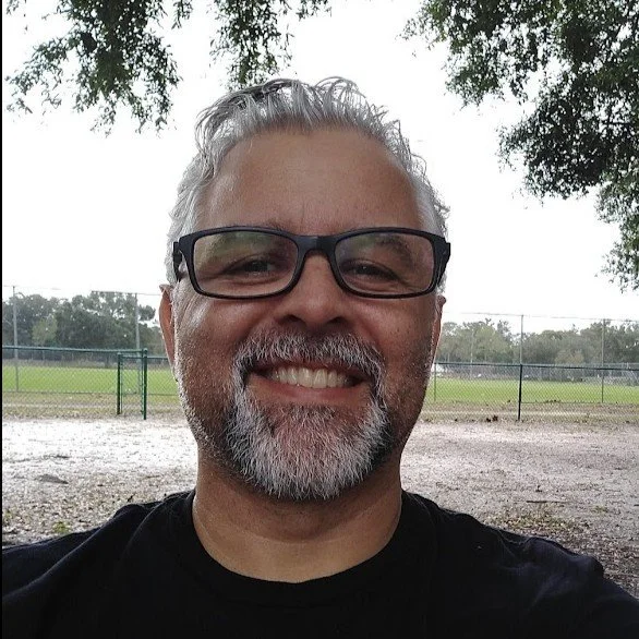 A smiling middle-aged man with glasses, gray hair, and a beard, outdoors on a cloudy day in a park or playground with trees and a fence in the background.