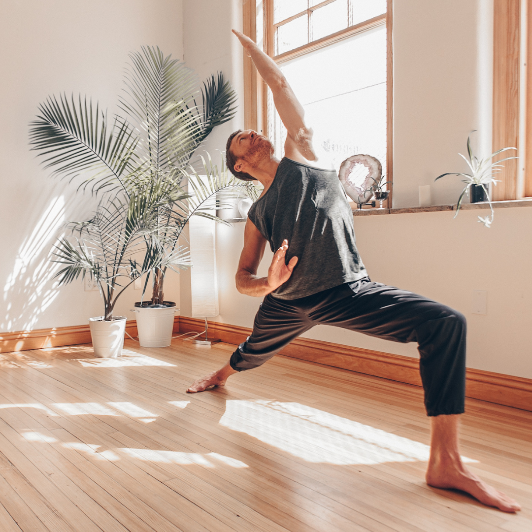 A shirtless man with short brown hair and blue eyes standing against a plain white background, looking directly at the camera with hands pressed together in a gesture of prayer or meditation.