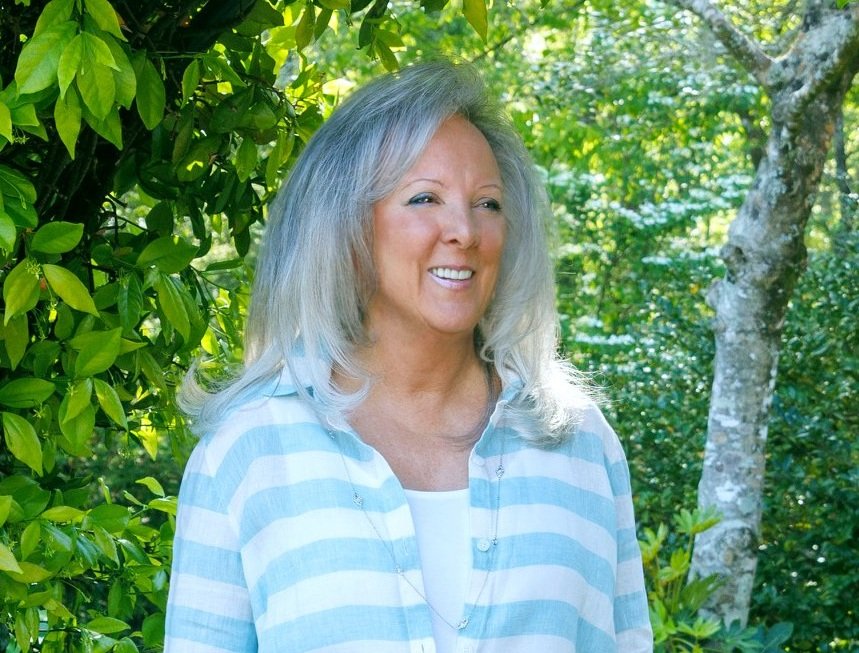 A middle-aged woman with long gray hair, smiling, standing outdoors in front of green bushes and trees, wearing a light blue and white striped shirt.