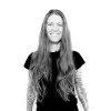 Black and white photo of a person with long hair and tattoos, wearing a black T-shirt, smiling against a plain background.