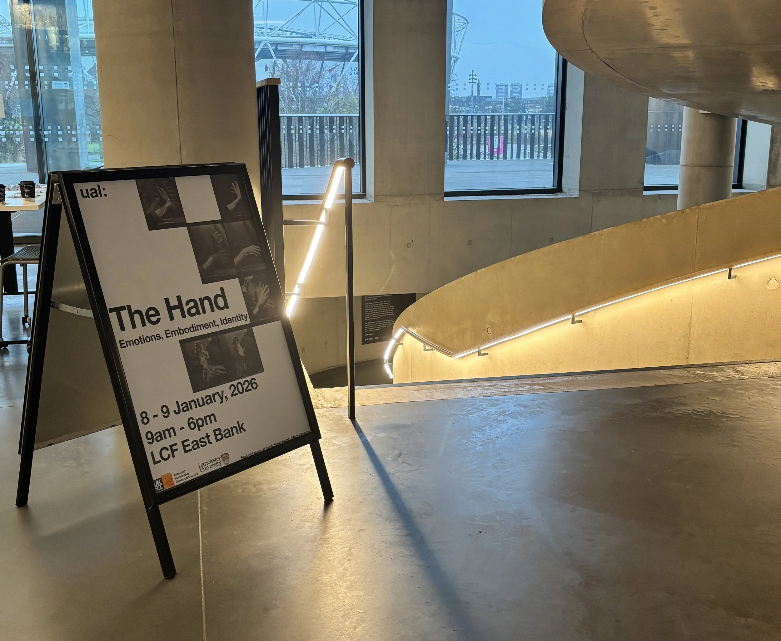 A poster on an easel advertises "The Hand" conference at LCF East Bank, set near a curved, well-lit staircase.
