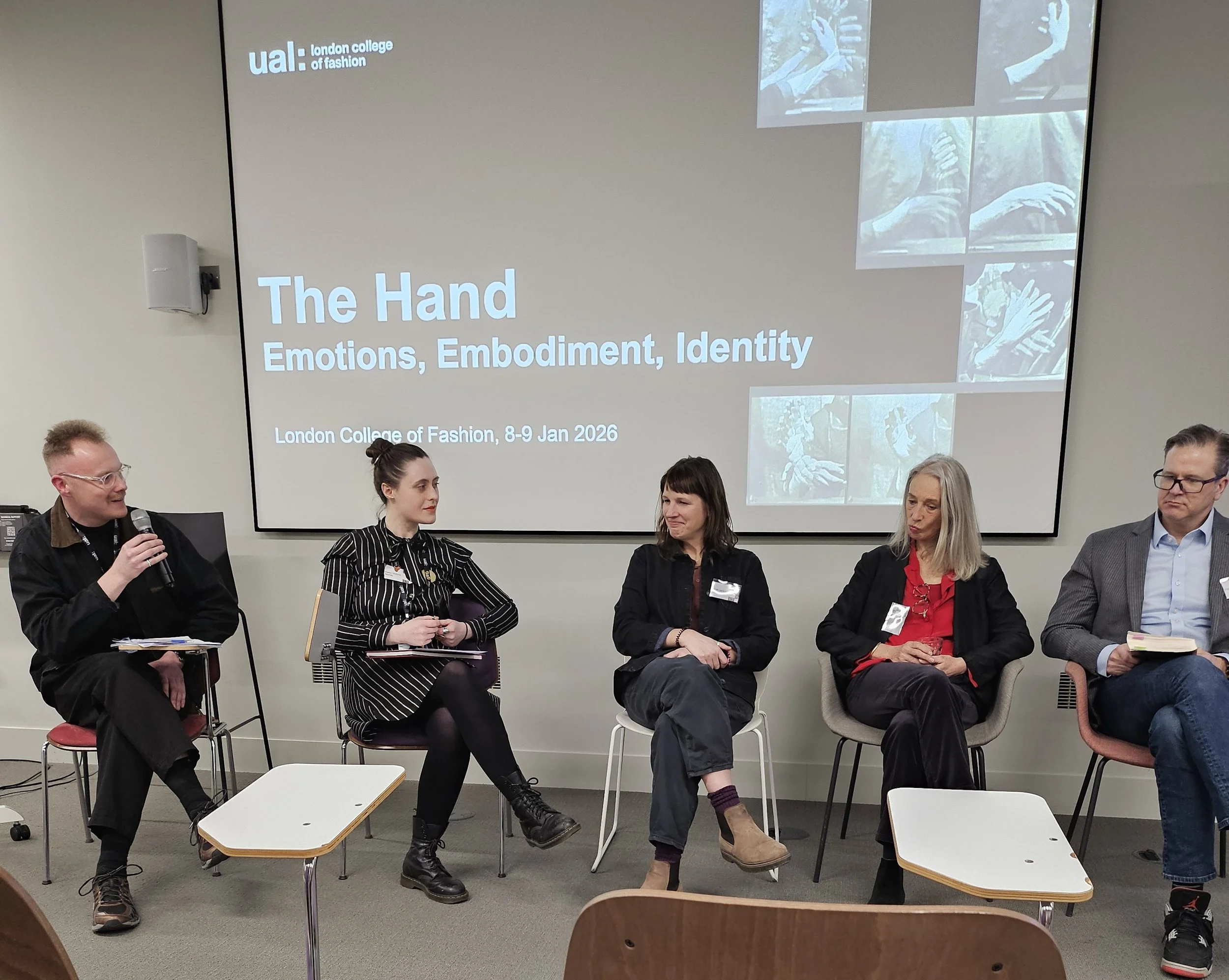 A panel of five people sits in discussion, engaging in conversation. Behind them, a slide reads "The Hand: Emotions, Embodiment, Identity" at the London College of Fashion event in 2026.