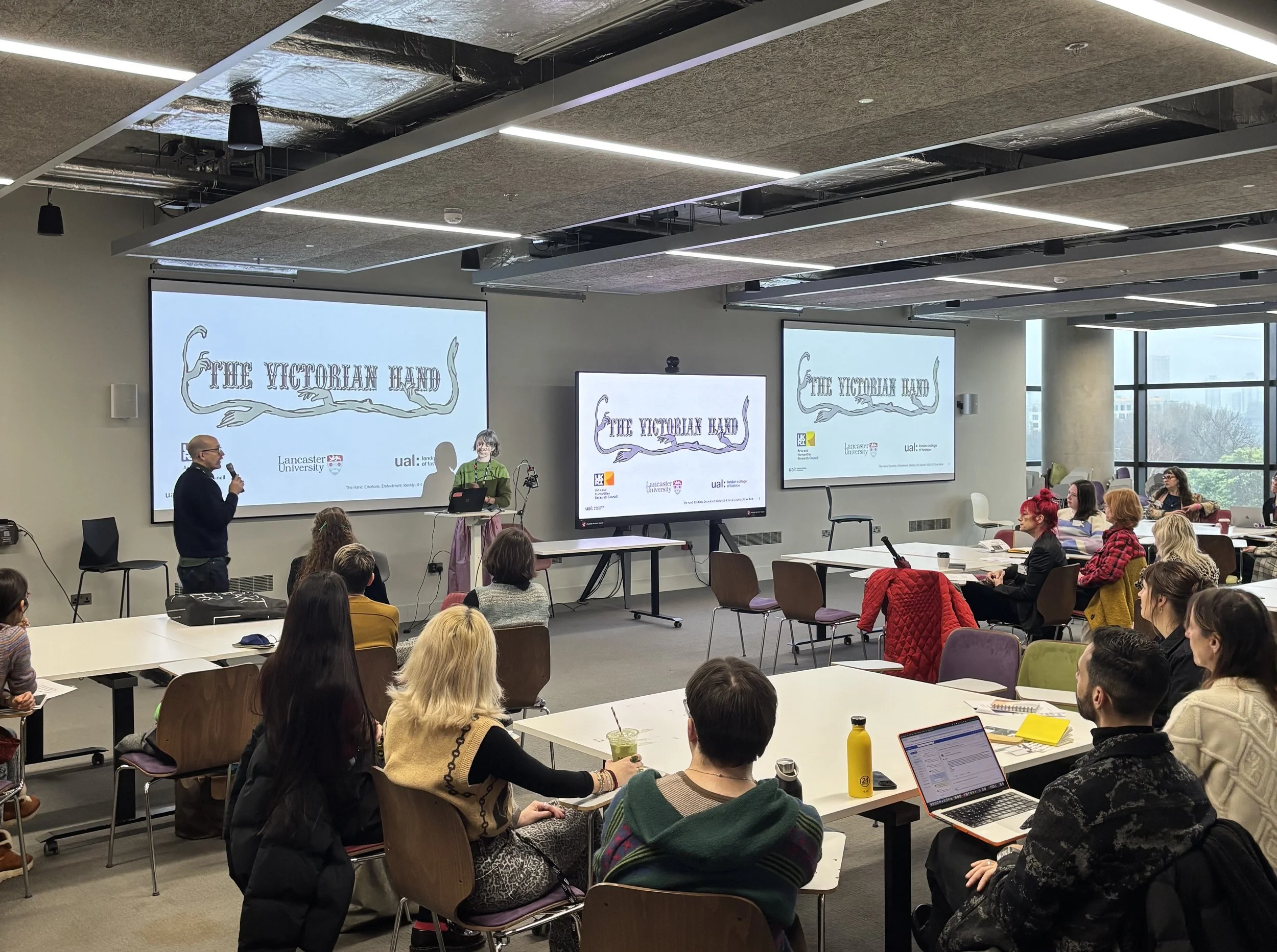 A conference room with people seated at tables, attentively watching a presentation. Two large screens display The Victorian Hand logo.