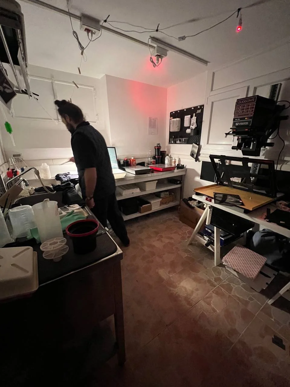 Camera oscura con un uomo che lavora su un tavolo industriale, attrezzature di stampa analogica e strumenti vari, pareti bianche, luce rossa, pavimento in piastrelle, ambientazione di un laboratorio o studio tecnico.
