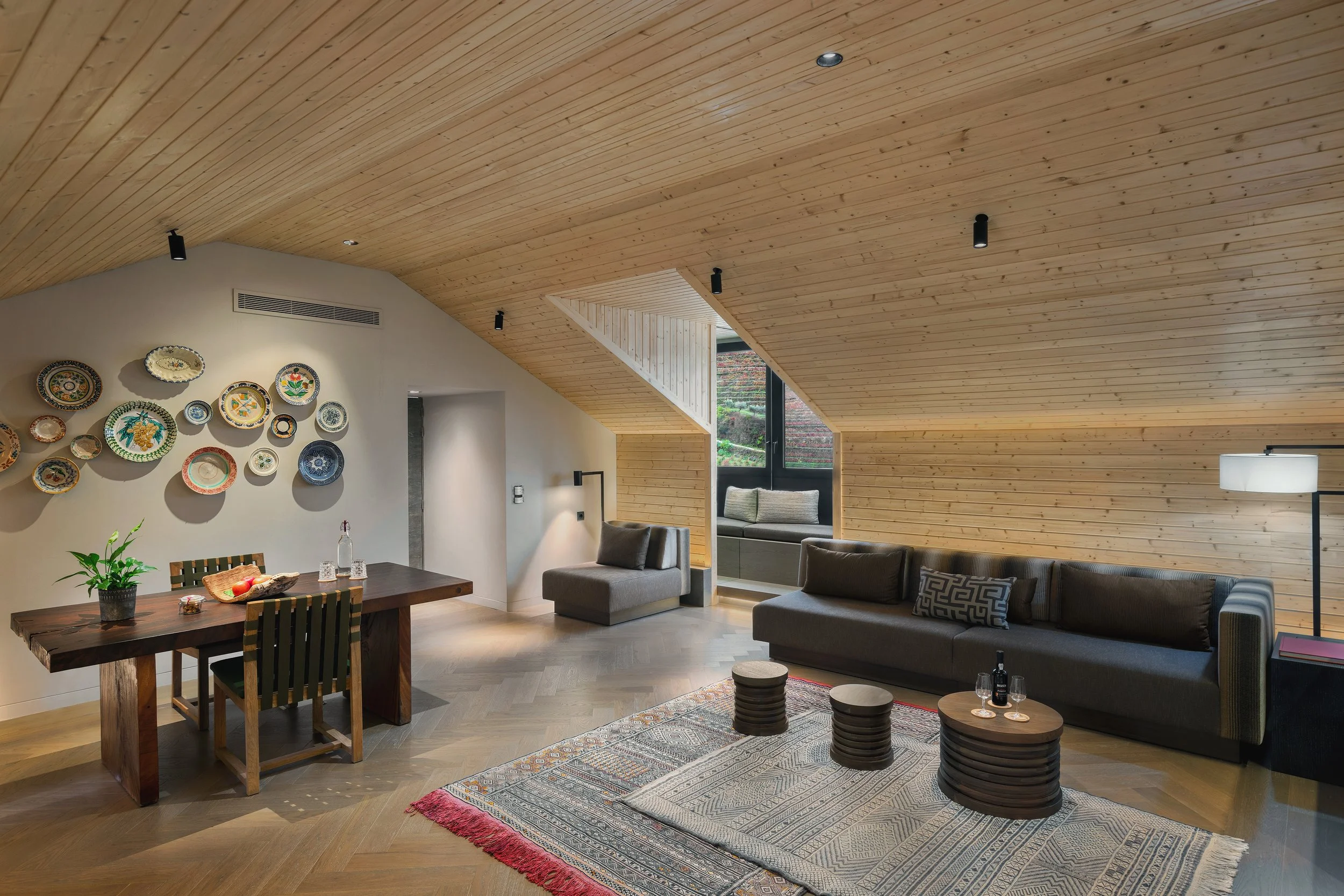 Living room with wooden sloped ceiling, a large gray sofa, a wooden coffee table, a rug, a small side table, and a window seat with cushions, decorated with colorful plates on the wall.