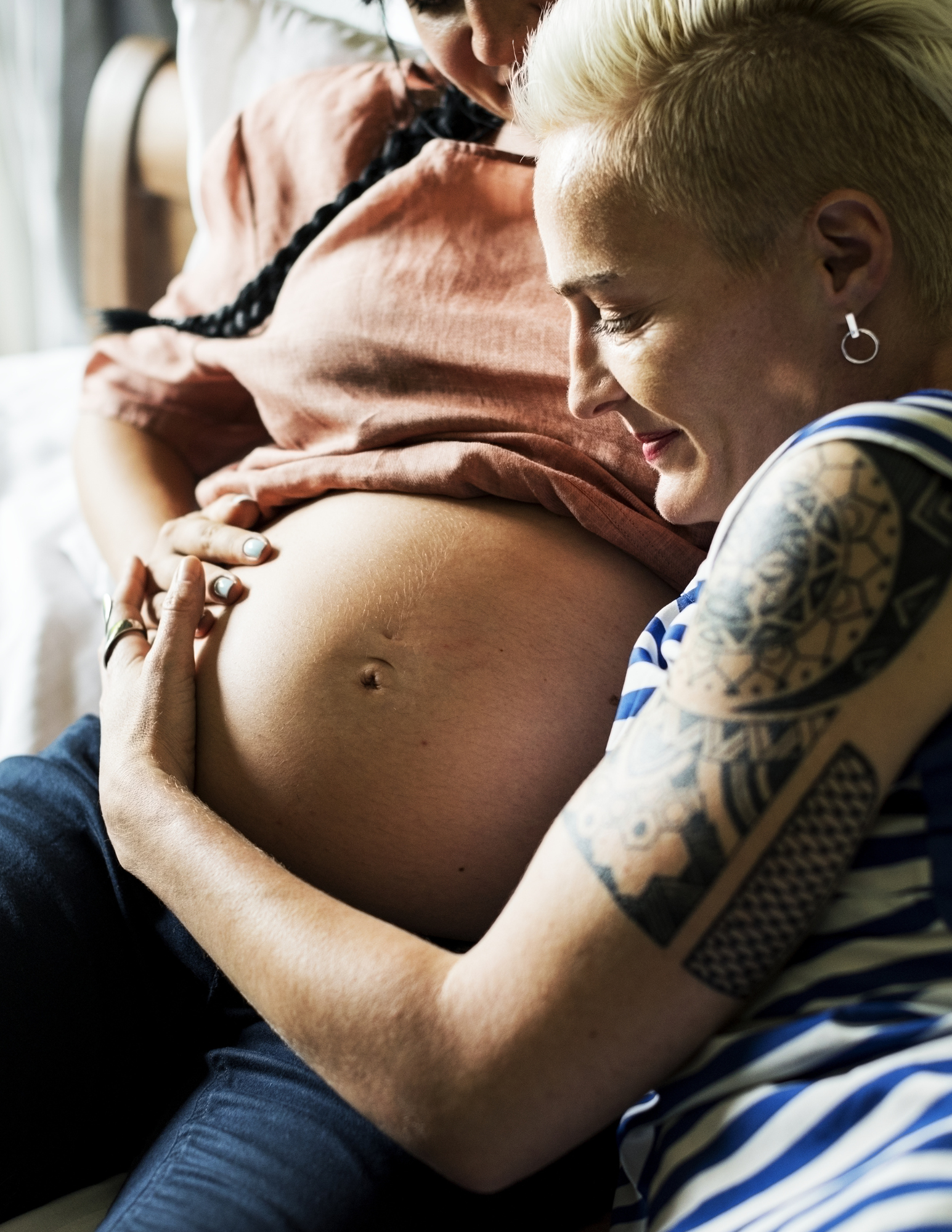 A person with tattoos on their arm gently touching a pregnant woman's belly, both smiling and engaging in a moment of intimacy.