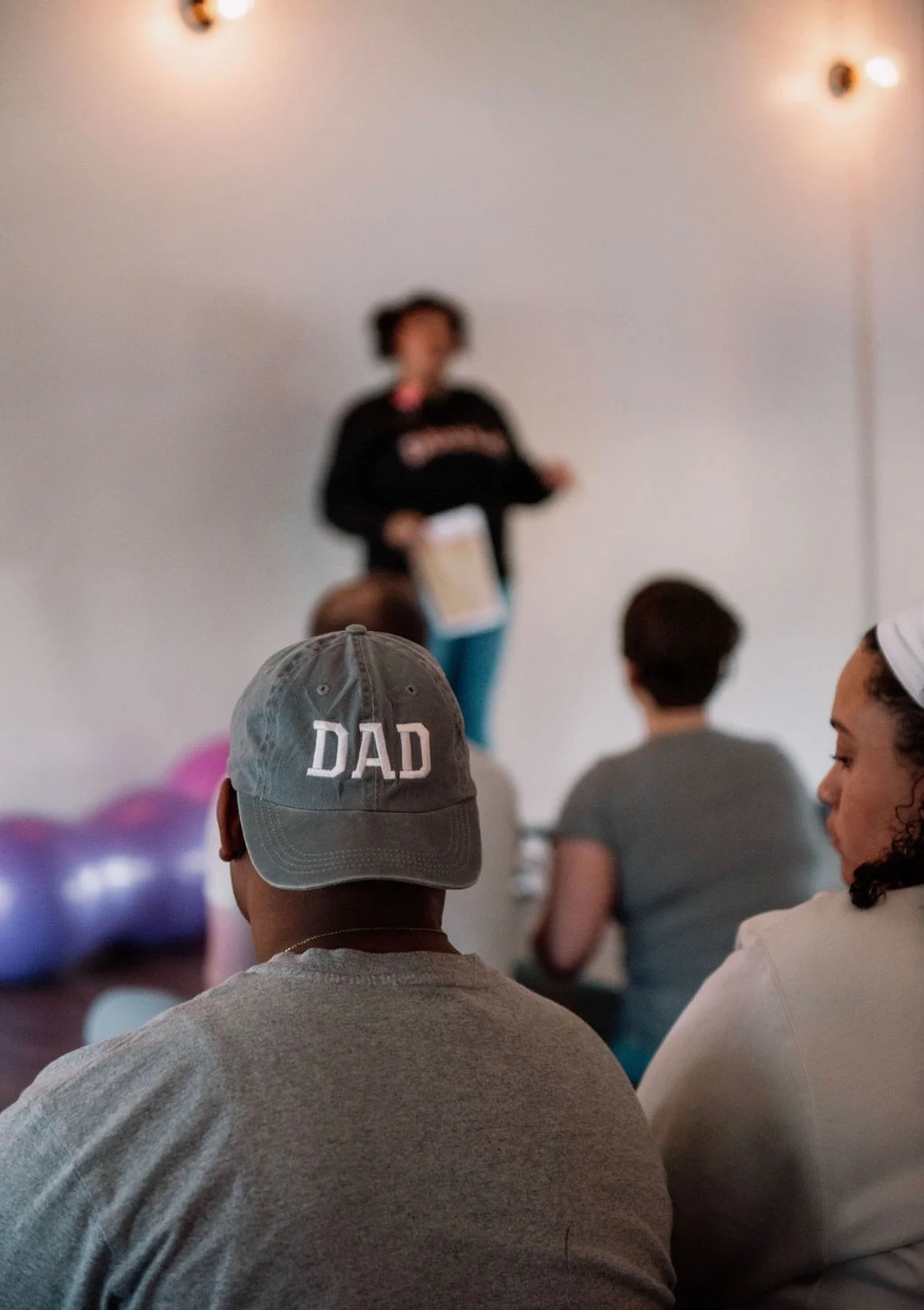 Audience listening to a speaker at a presentation, with a person wearing a gray cap labeled 'DAD' in the foreground.