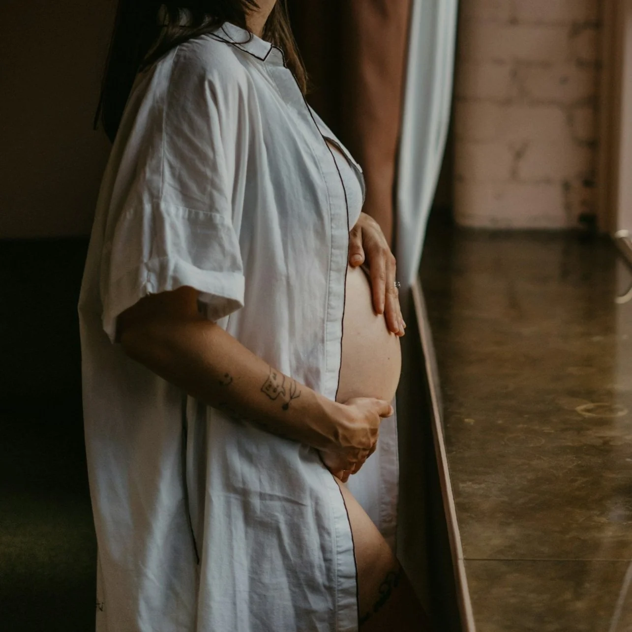 A woman in a white shirt holds her pregnant belly, standing near a mirror with a rustic wooden frame inside a dimly lit room.