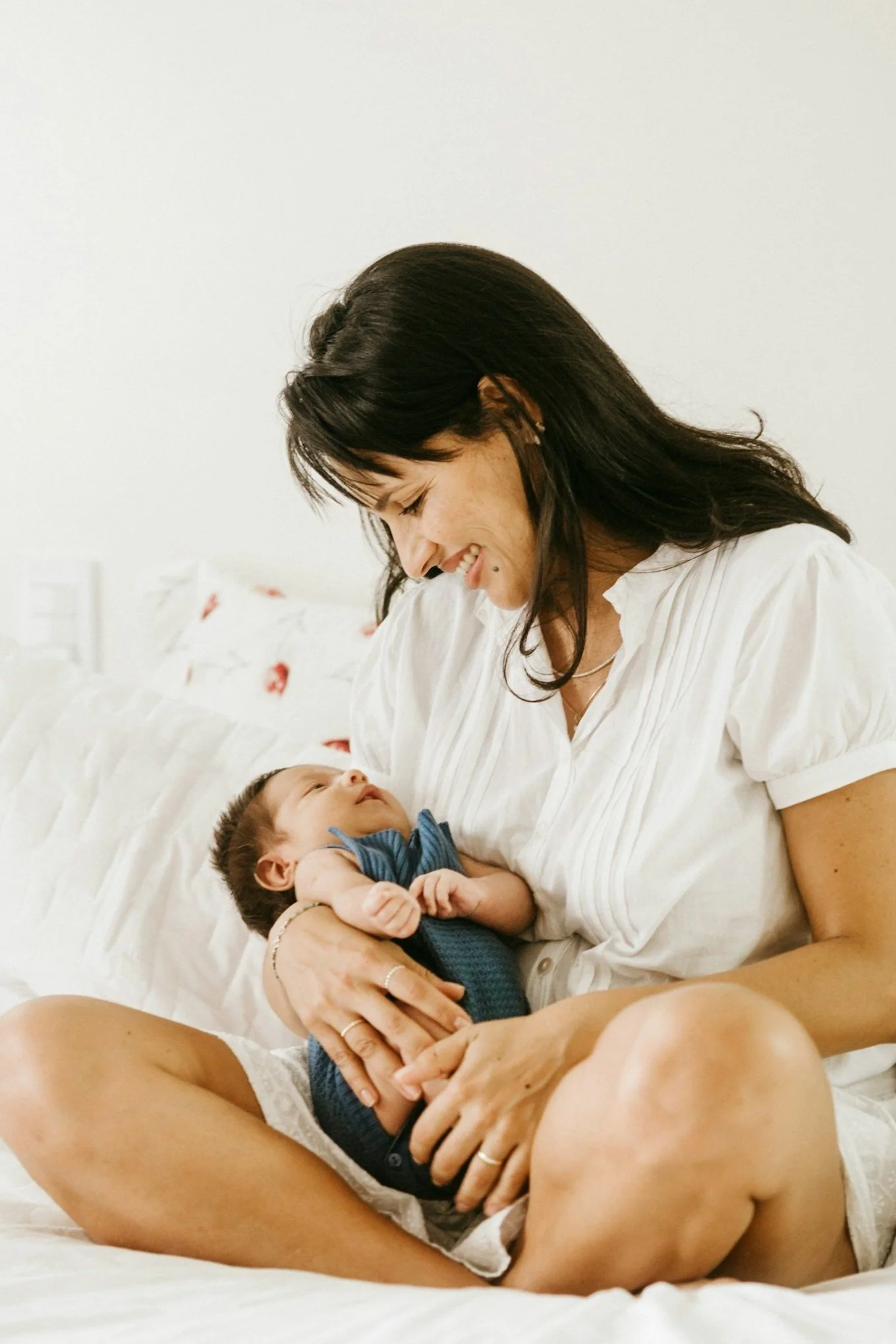 A woman with dark hair smiling and looking at a baby she is holding and sitting on a bed.