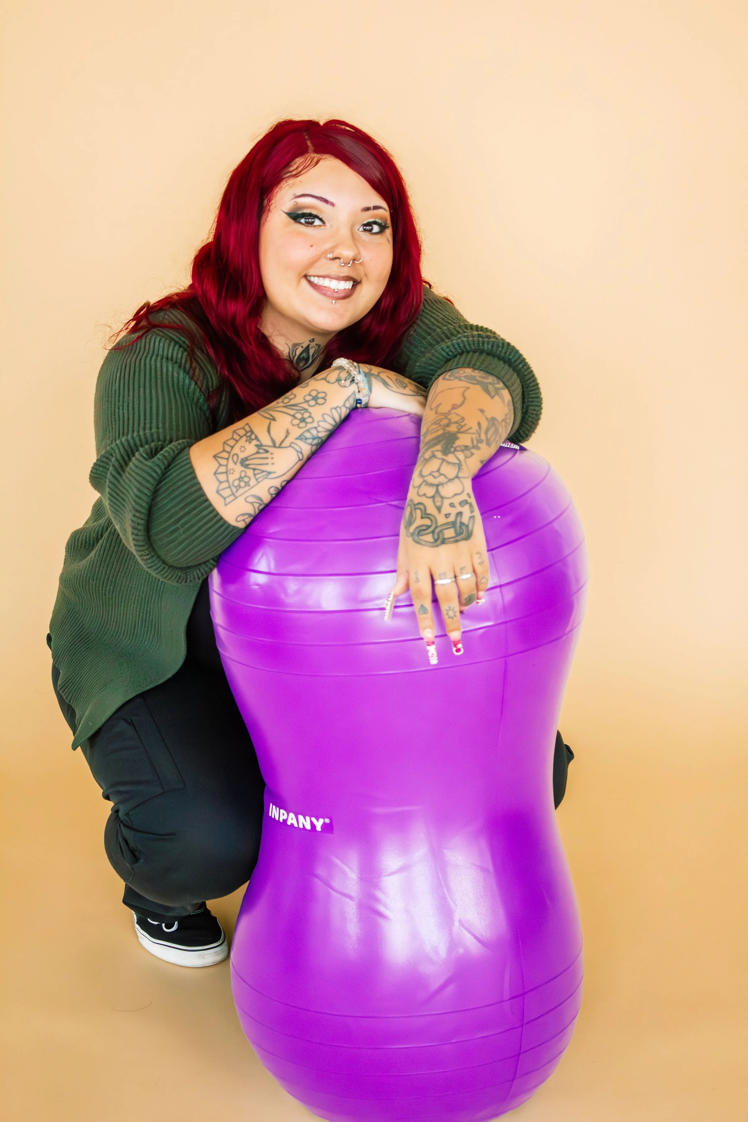 A woman with red hair and tattoos, smiling and squatting next to a large purple exercise ball against a beige background.