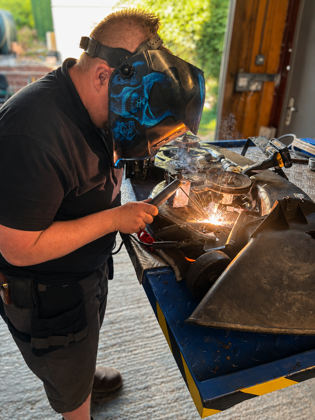 Person welding metal parts at workbench, wearing a welding helmet, using a welding torch, sparks and smoke visible, workshop setting with tools and equipment.