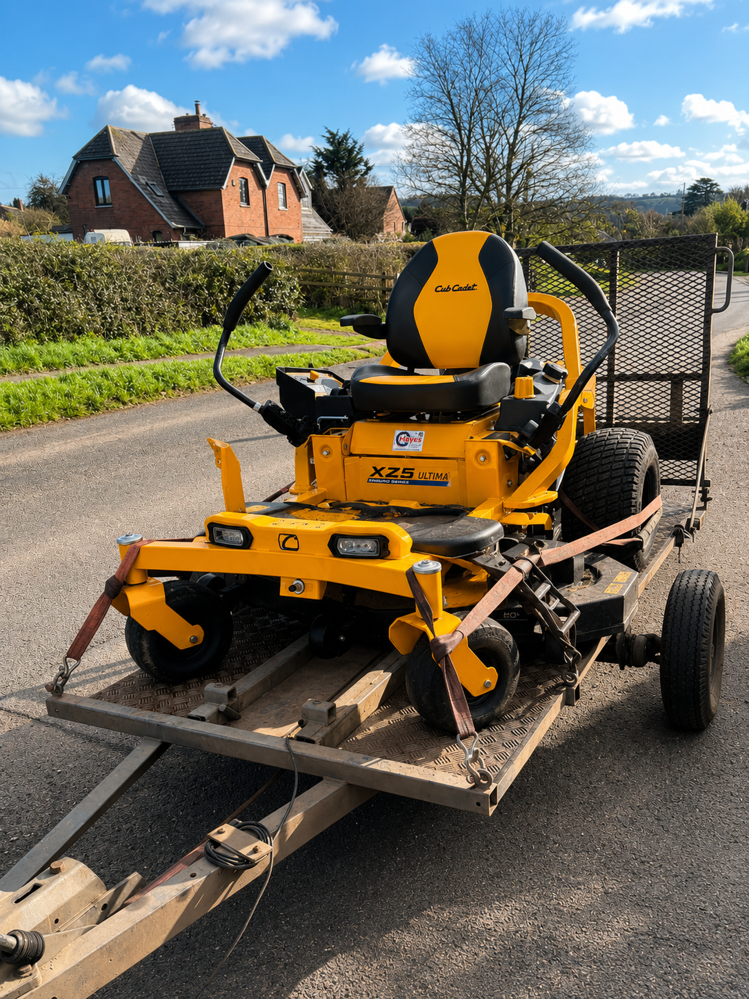A yellow commercial mower with a black seat parked on a trailer on a suburban street with houses and trees in the background.