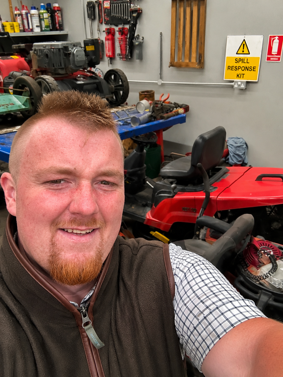 A man taking a selfie in a workshop with various tools, a ride-on lawnmower, and safety signs on the wall.