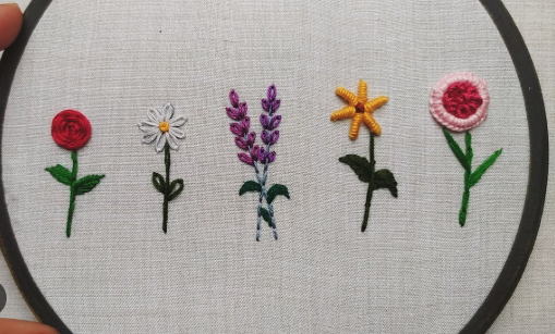 Embroidery hoop with five embroidered flowers: a red rose, a daisy, lavender, a yellow starflower, and a pink flower.