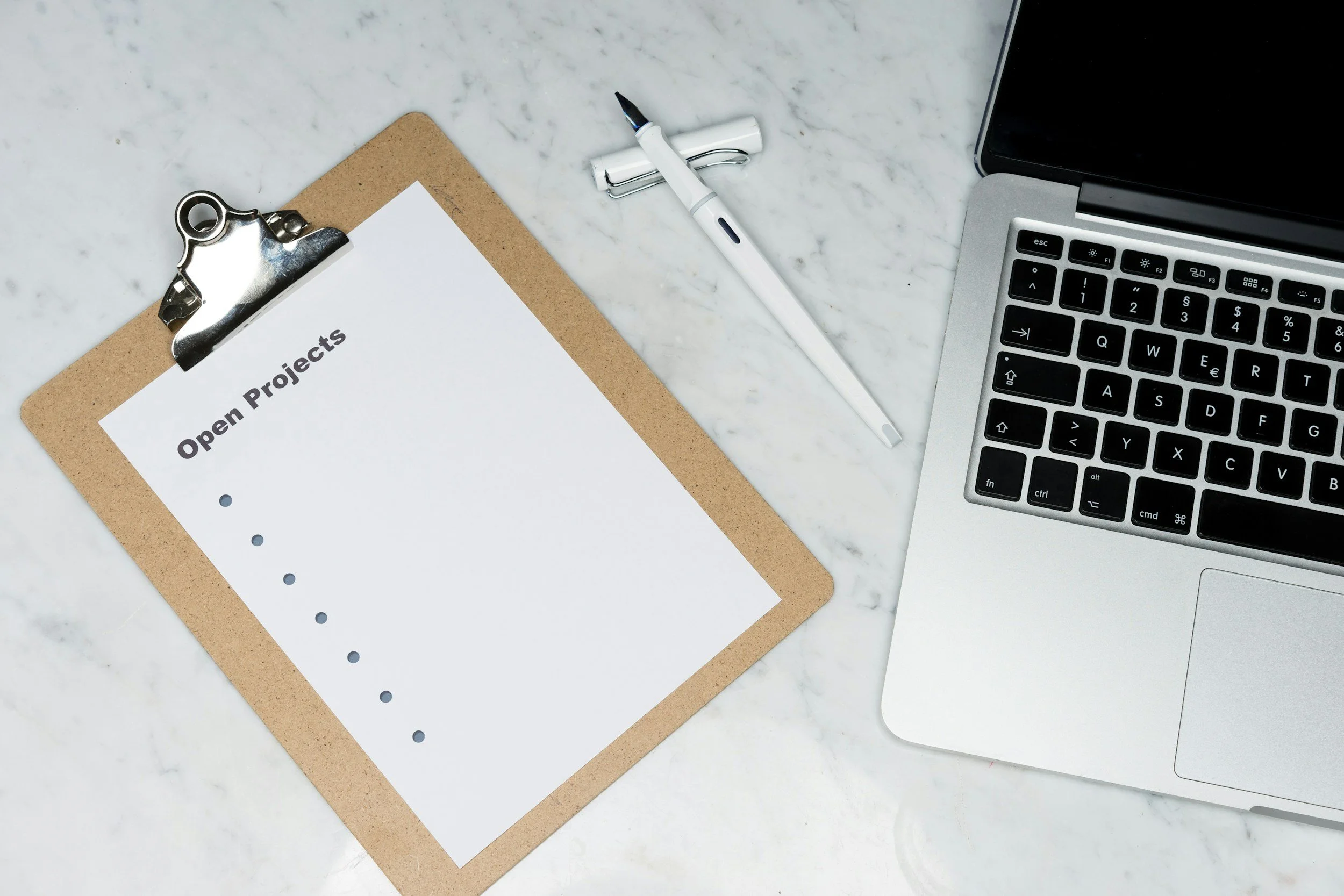 Top view of a clipboard with a sheet titled 'Open Projects', a pen, and a laptop on a marble surface.
