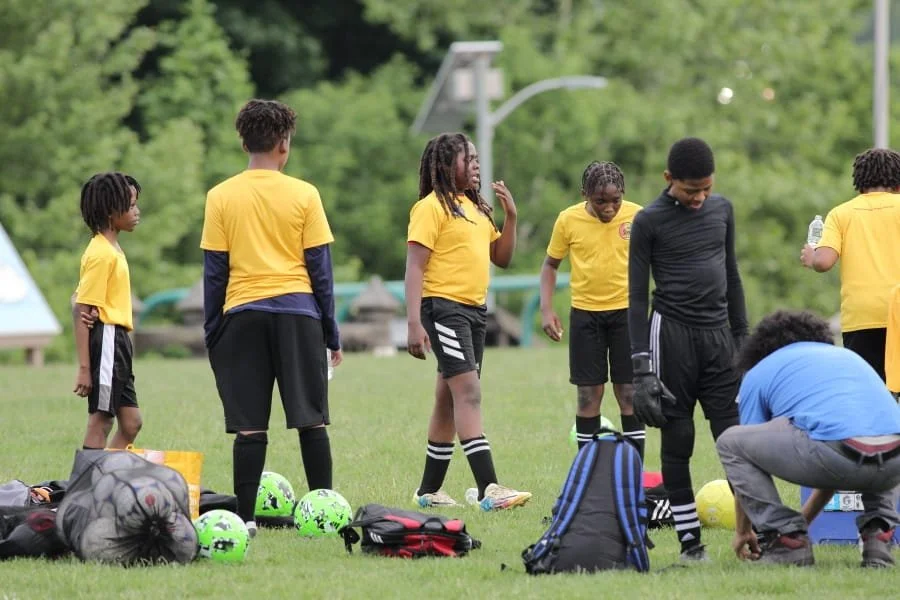 A group of young soccer players wearing yellow jerseys standing on a grassy field with various bags and soccer balls around them.
