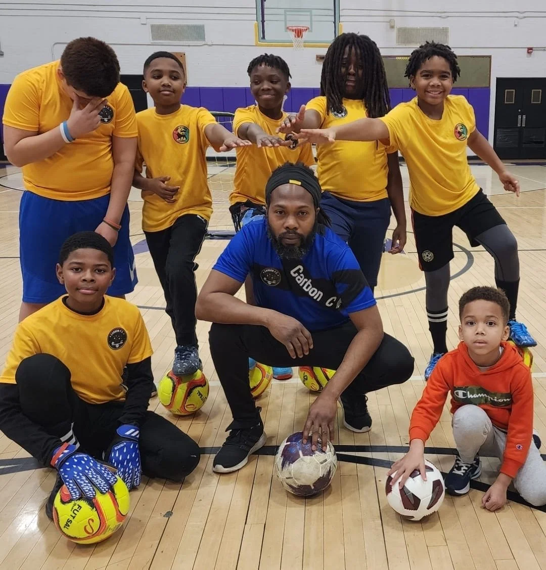 Group of kids and a coach in a gym, wearing sports uniforms, posing with soccer balls.