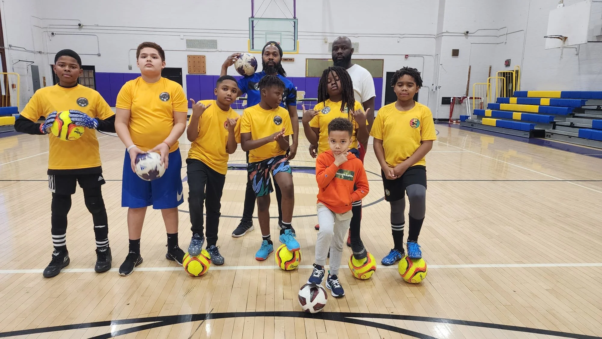 Group of children and two adults on a gymnasium floor holding soccer balls, wearing yellow shirts and athletic gear.