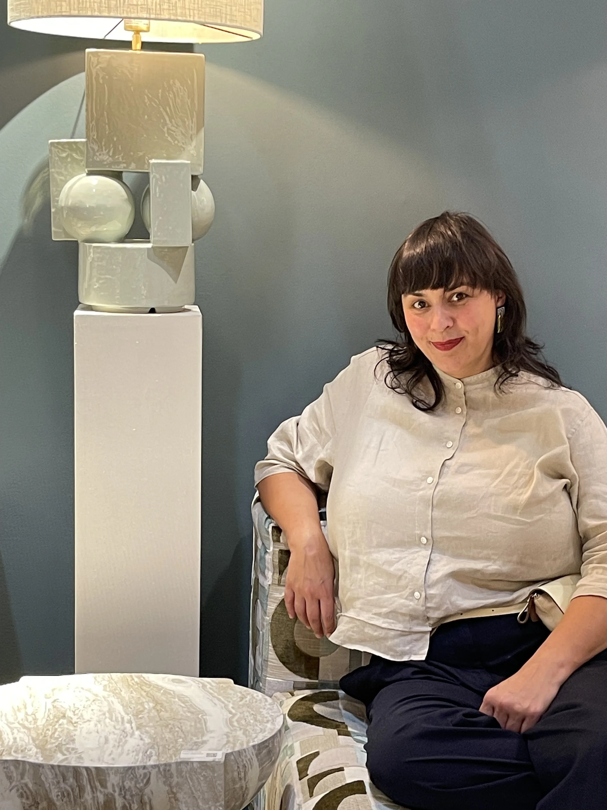 Een vrouw met bruin haar en een witte blouse zit op een stoel, naast een ronde marmeren tafel en een moderne lamp tegen een blauwgrijze muur.