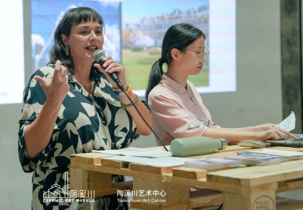 Inge Simonis geeft een lezing in Jingdezhen, zij spreekt in een microfoon en de vertaler kijkt naar een stuk papier. Op de achtergrond een scherm met een afbeelding en tekst in het Chinees.