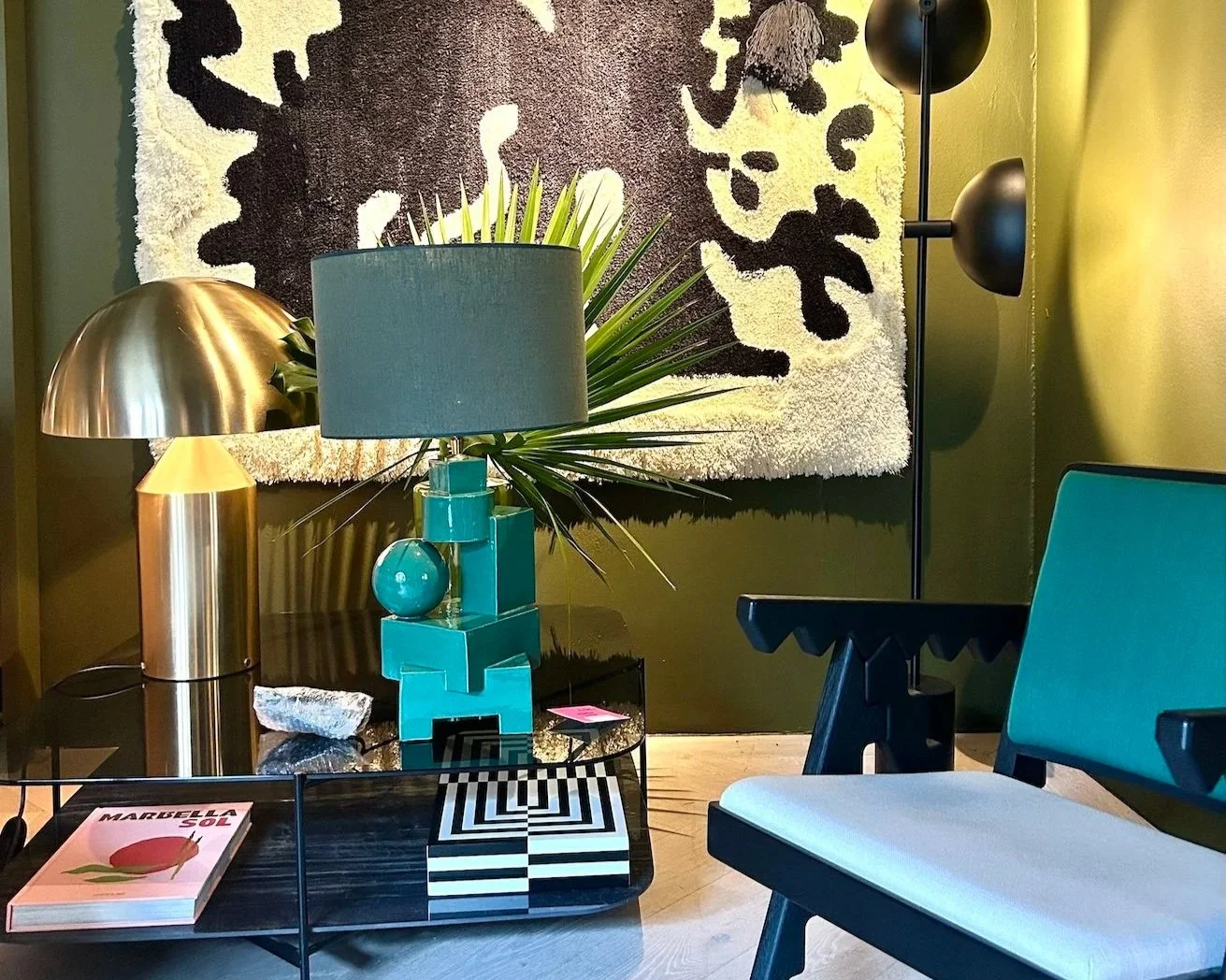 Een modern interieur met een zwarte salontafel, een groene keramische tafellamp van Inge Simonis een groene muur, een muurhanger met zwart en wit patroon, en een groene stoel.