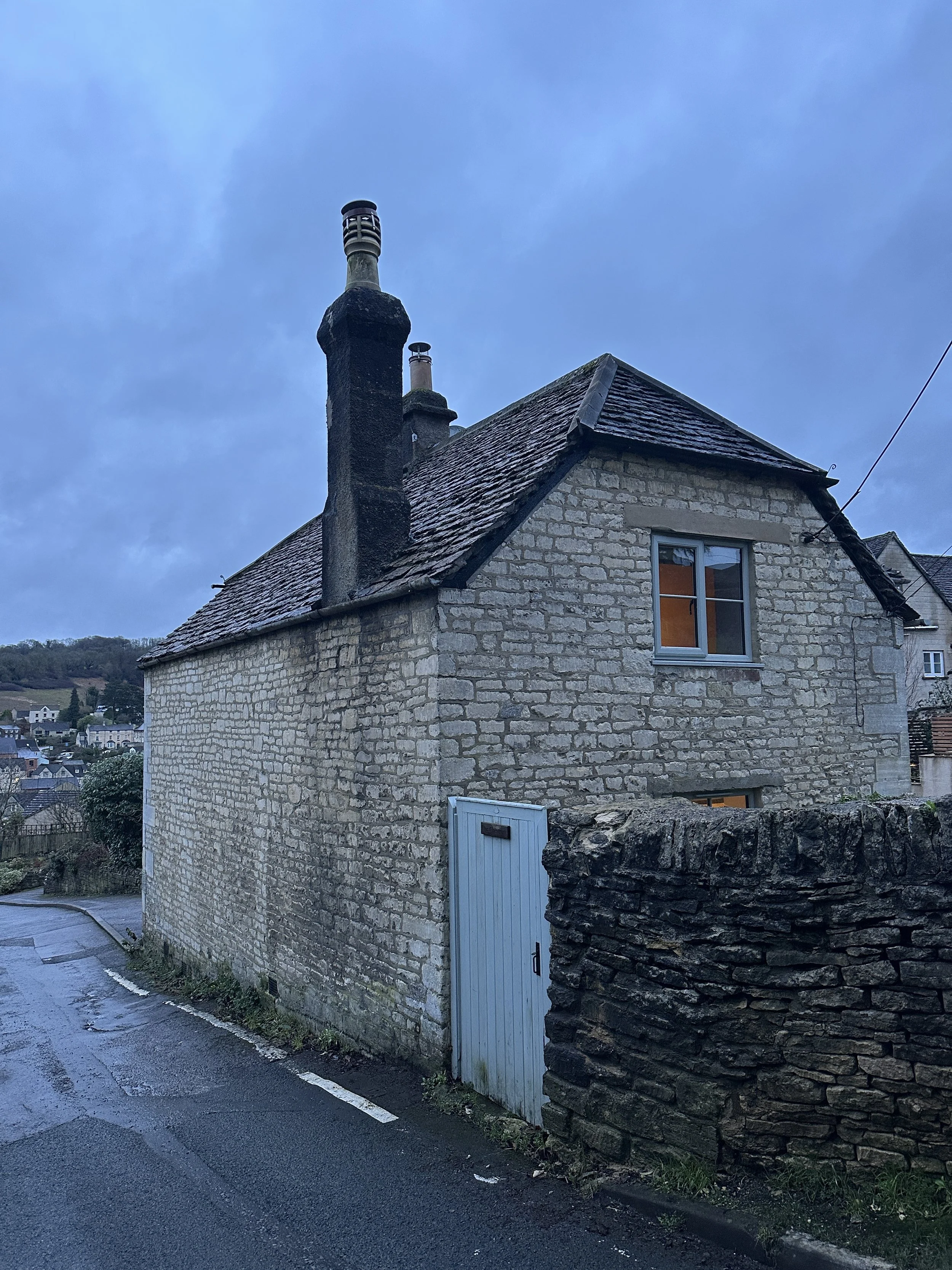 A stone building with a sloped roof and a window, situated on a street corner with a stone wall and a metal gate. Two chimneys are visible on the roof against a blue sky with some clouds.
