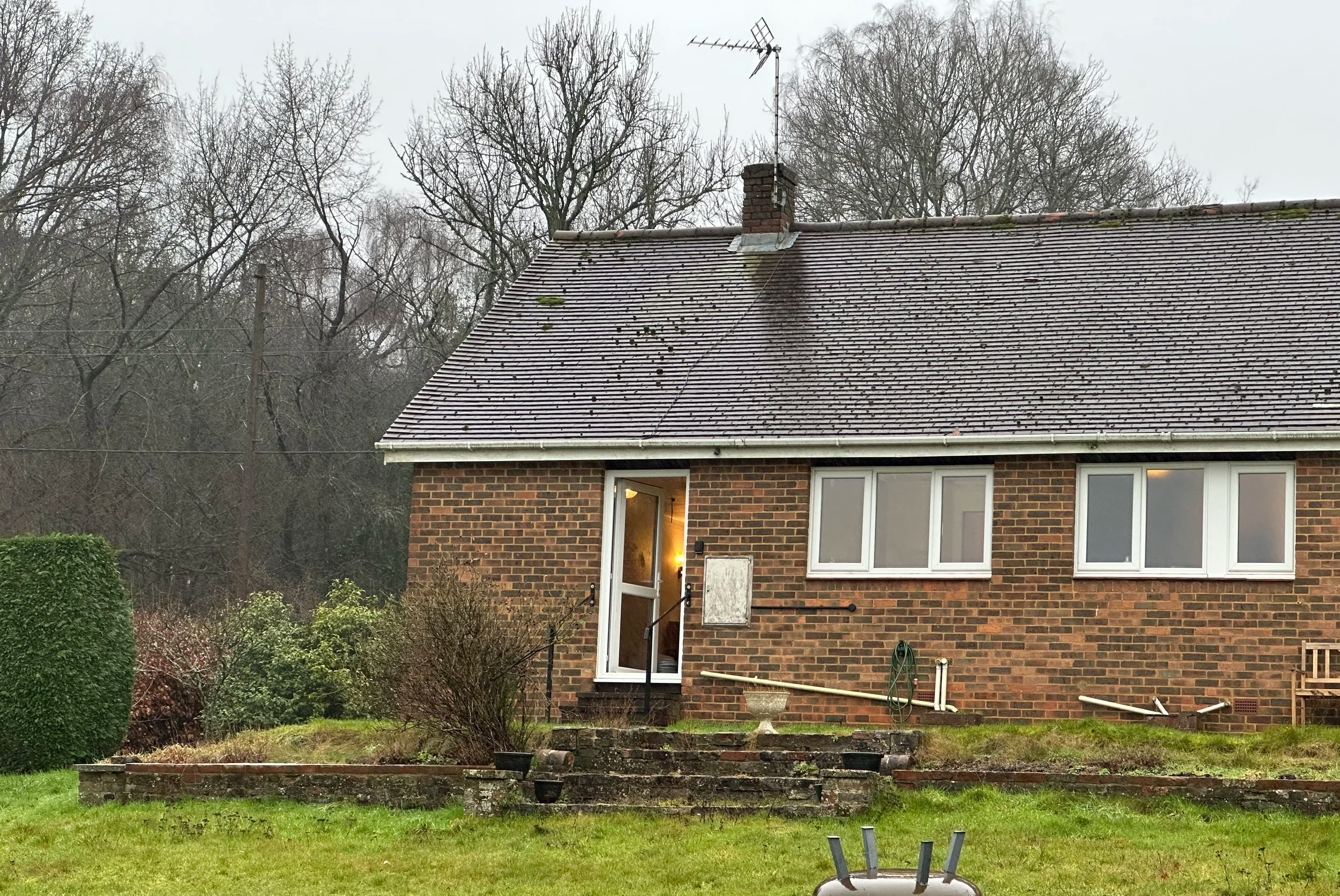 A brick bungalow with a tiled roof, chimney, and television antenna, surrounded by trees and a green lawn with plants and outdoor equipment.