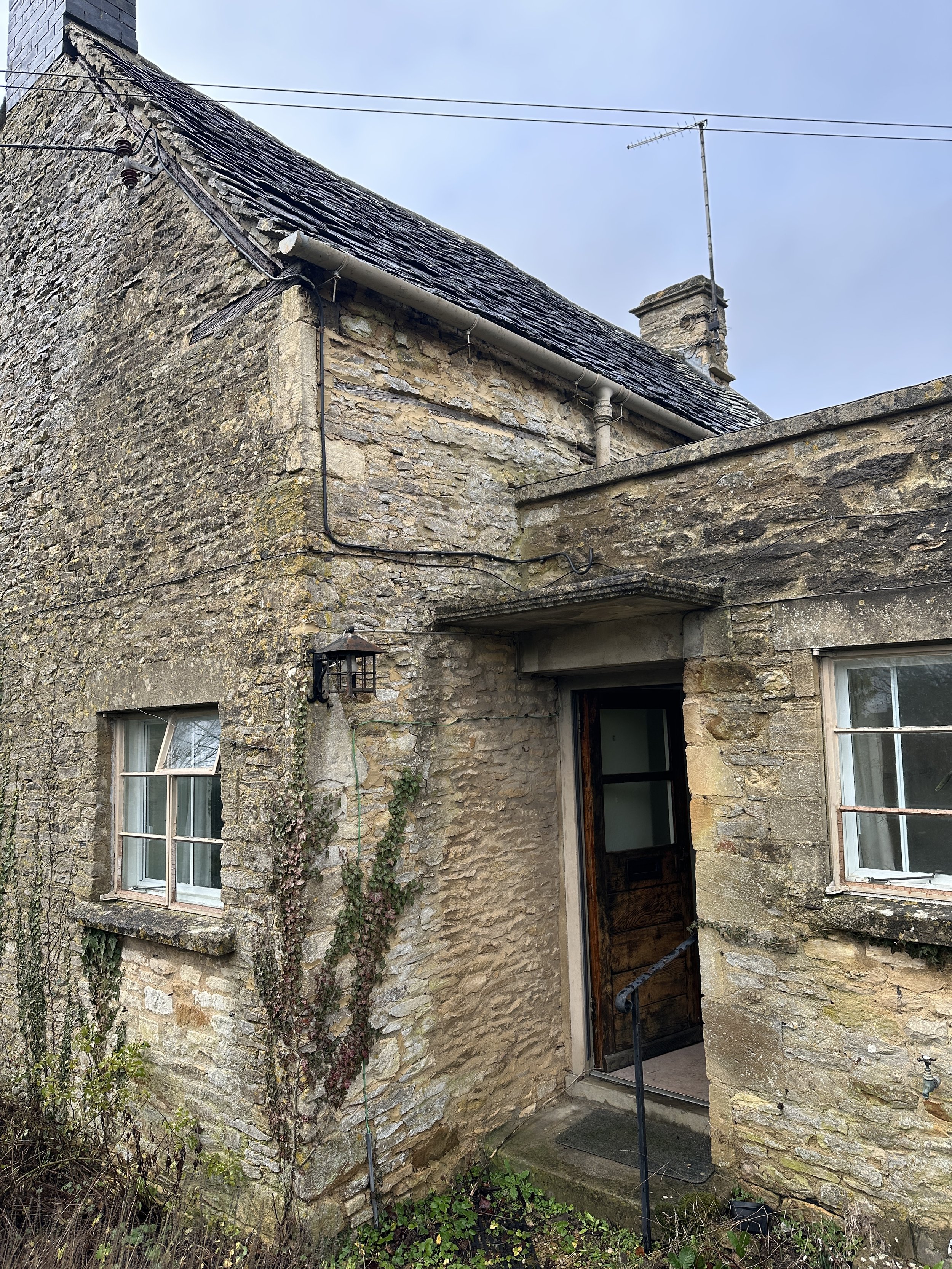 An old stone house with ivy growing on the wall, two windows, and a partially open door leading inside. The roof is covered with dark slates and there are some wires and an antenna on the roof.