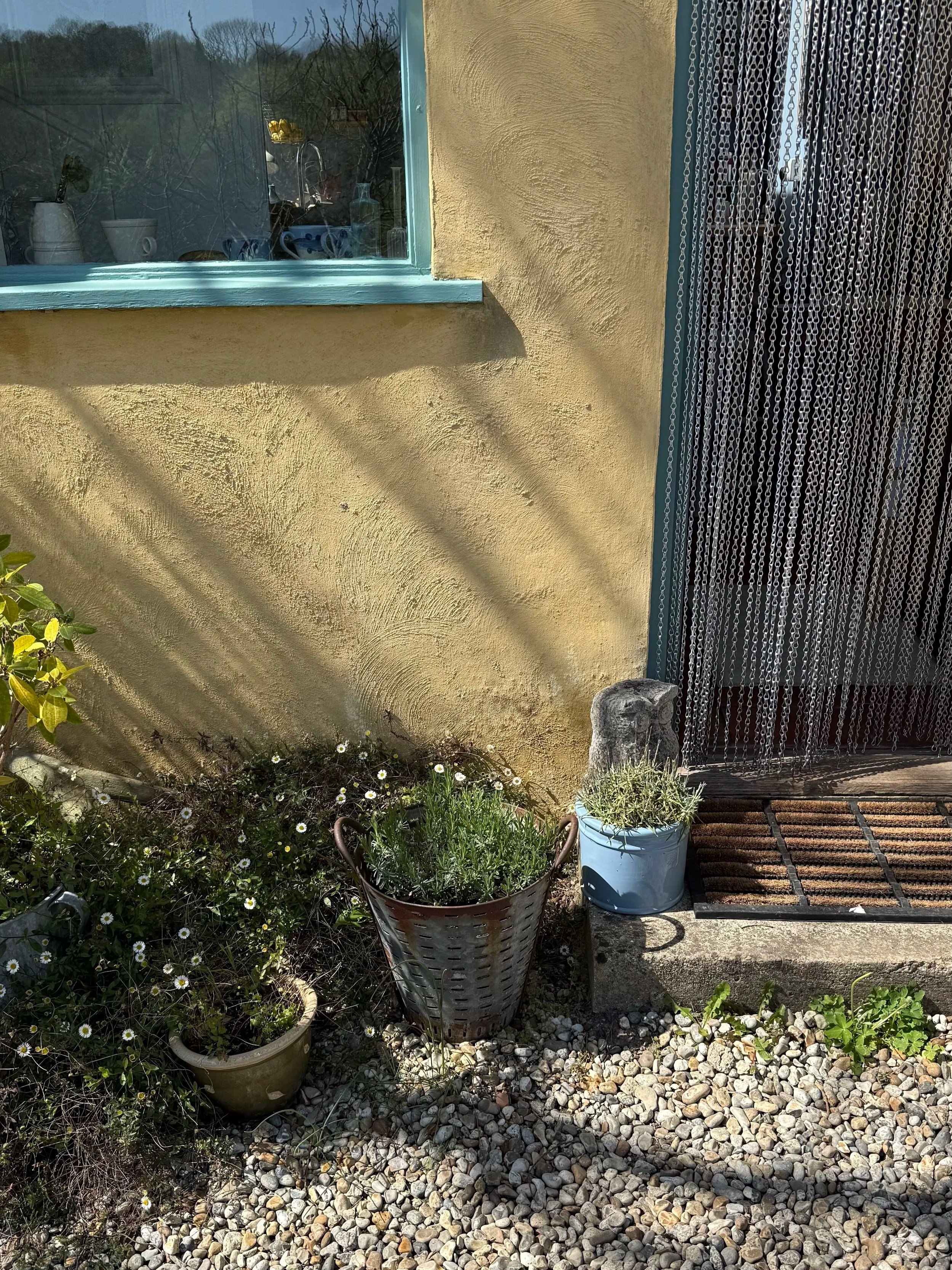 Sunlit porch area with a yellow exterior wall, a window with blue trim, potted plants including daisies, and a beaded curtain at the door. Gravel ground and some shrubbery are also visible.