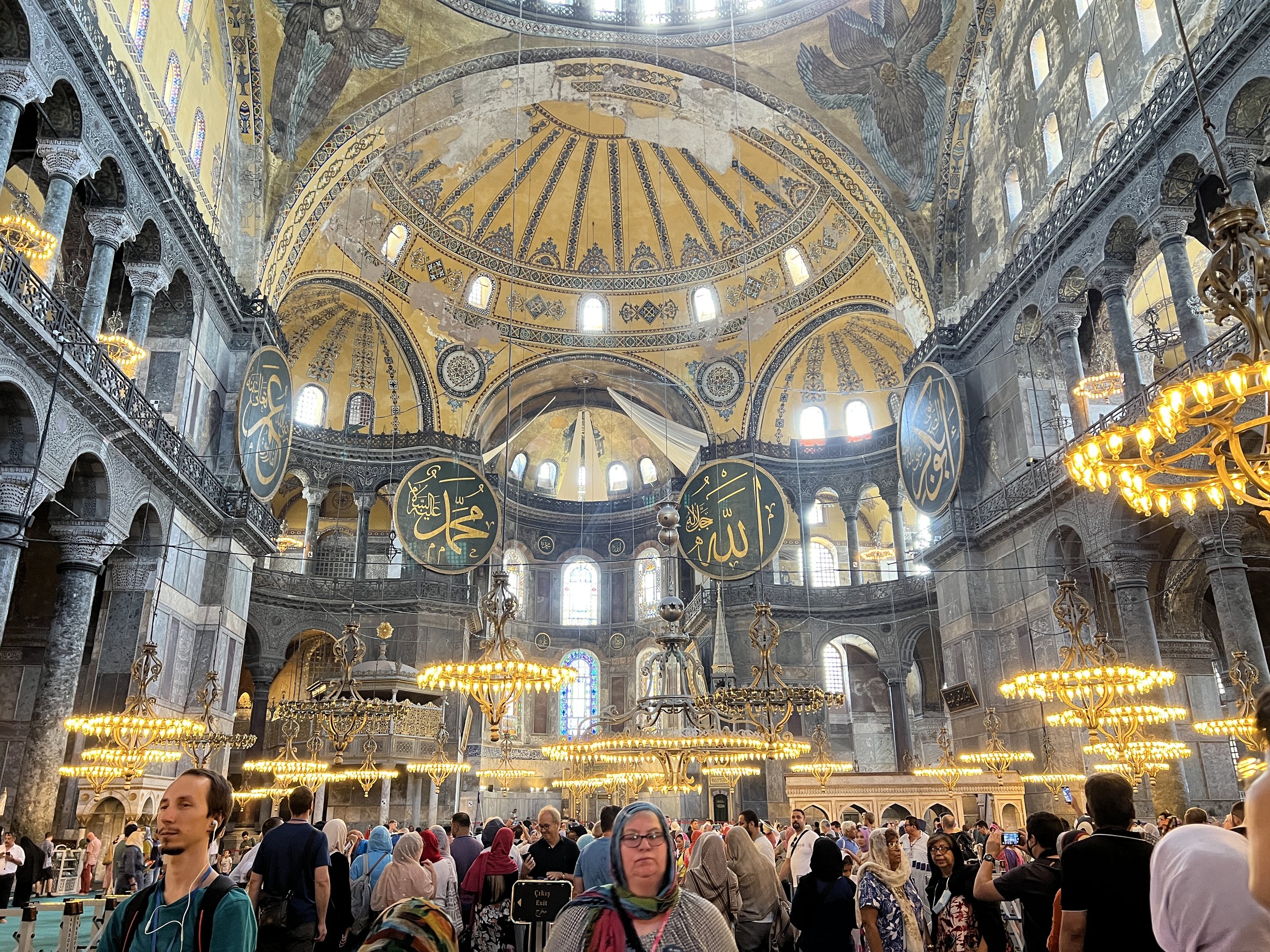 Interior of Hagia Sophia with ornate ceiling, large Islamic calligraphy plaques, and a crowd of visitors.