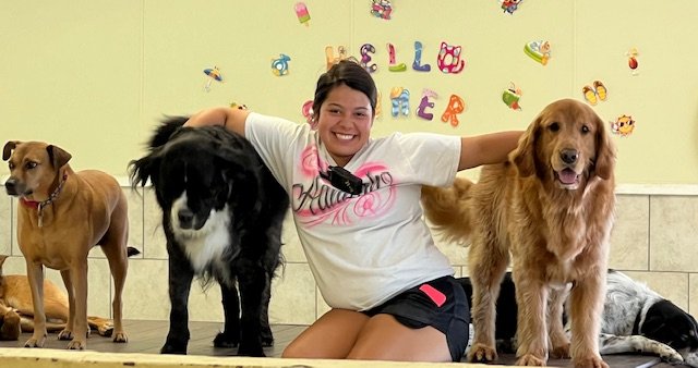 A woman smiling and posing with four dogs in front of a decorated wall that says "Happy Halloween" with colorful letters and Halloween-themed cutouts.