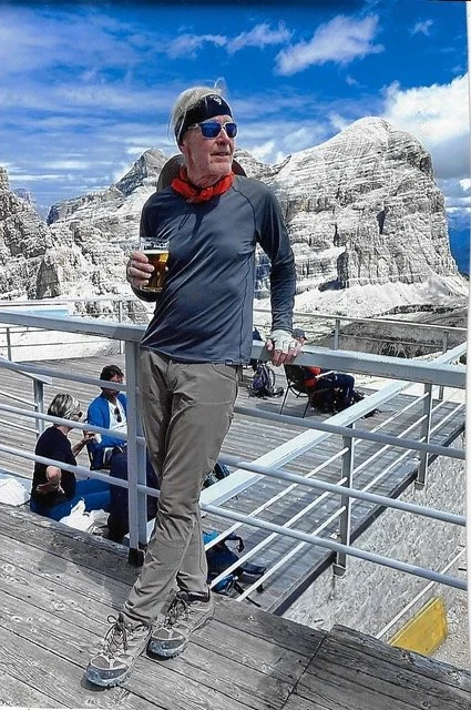 Man standing on a wooden viewing platform at a mountain site, holding a beer, wearing sunglasses, a headband, gray pants, and a long sleeve shirt, with rocky mountain formations in the background.