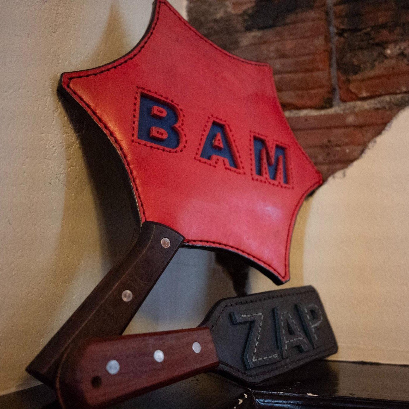 A large red leather paddle with the words BAM and a small leather paddle with the word ZAP are displayed in front of a wall that is white with a section of exposed brickwork