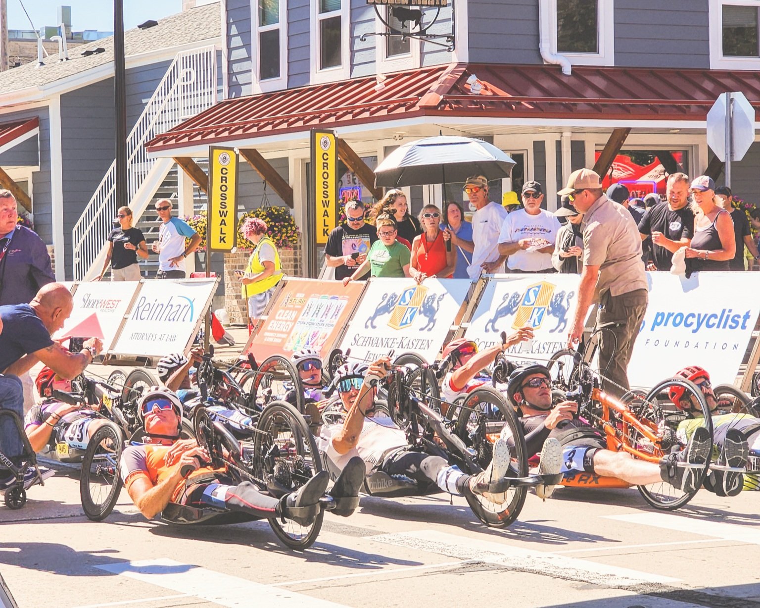 Tandem recumbent tricycle racers preparing at the starting line during a race event, with spectators watching from behind barricades on a sunny day.