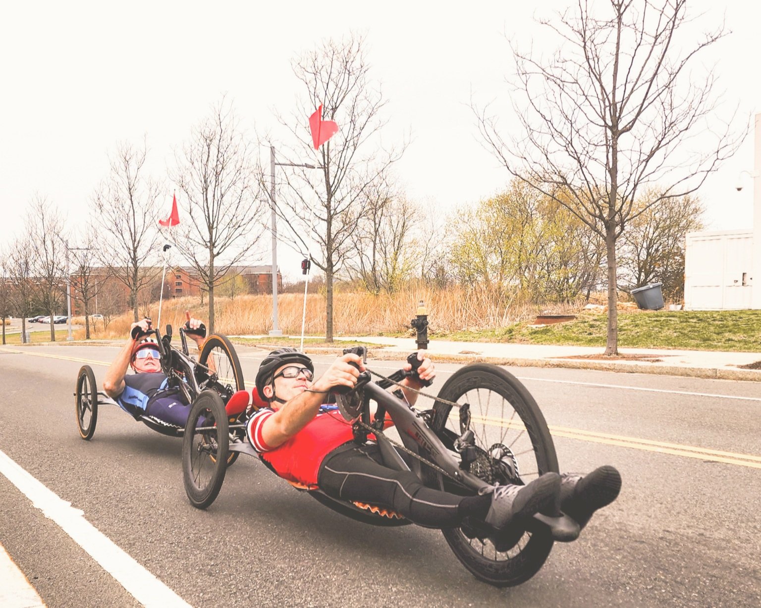Two people riding recumbent tricycles on a street, wearing helmets, with trees and a fire hydrant in the background.