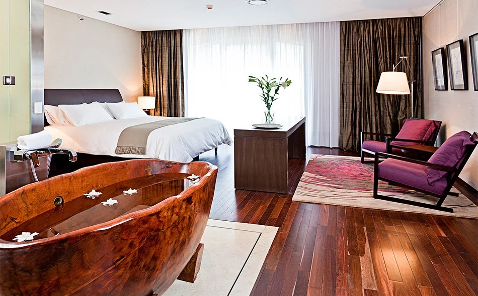 Luxury hotel room with a bathtub filled with water and flower petals, a bed with white linens, a wooden side table with a towel, a sitting area with two purple chairs, and large windows with curtains.