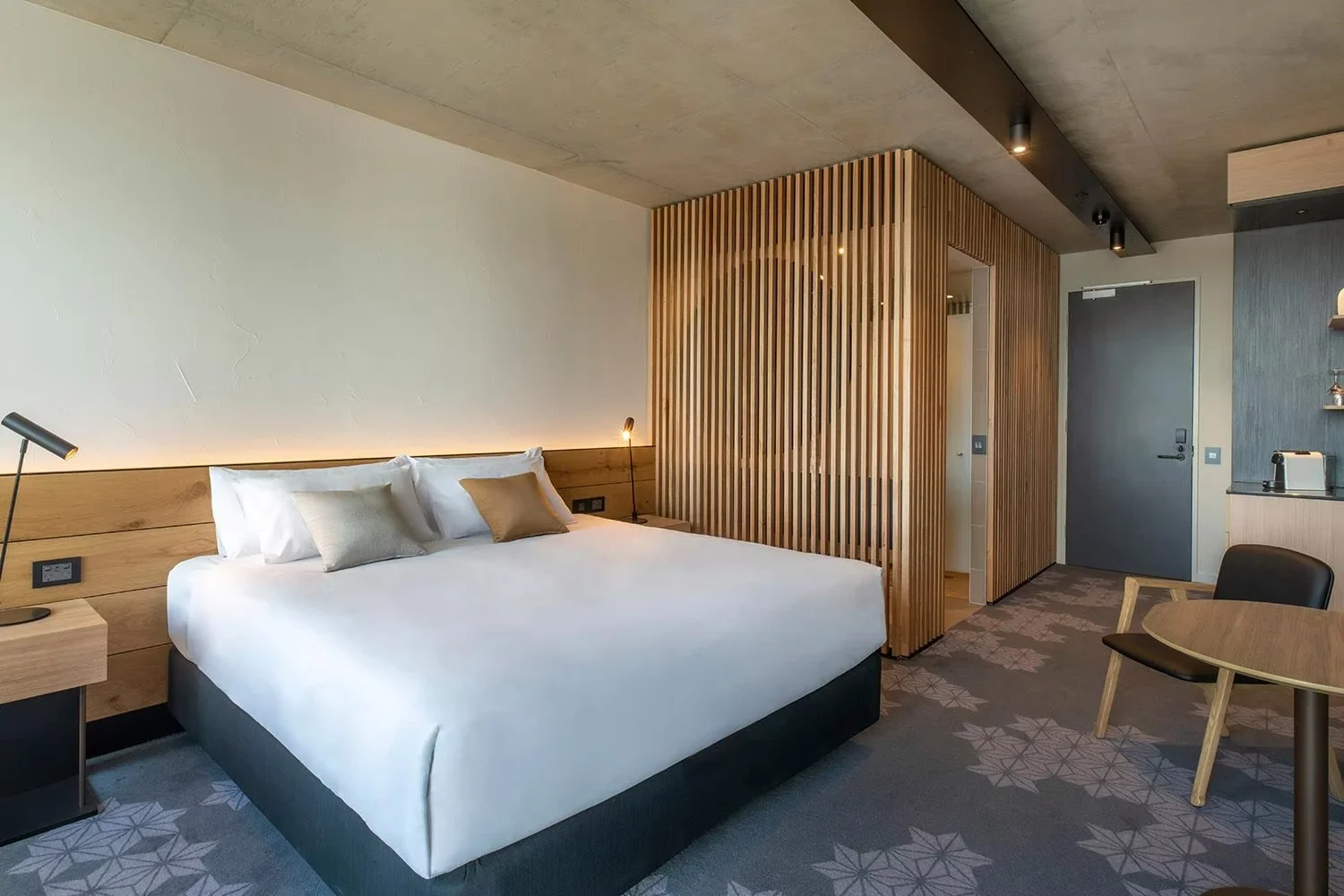 Modern hotel room with a large bed, wooden headboard, small side table with a lamp, wooden partition, table with chairs, and a kitchenette area.