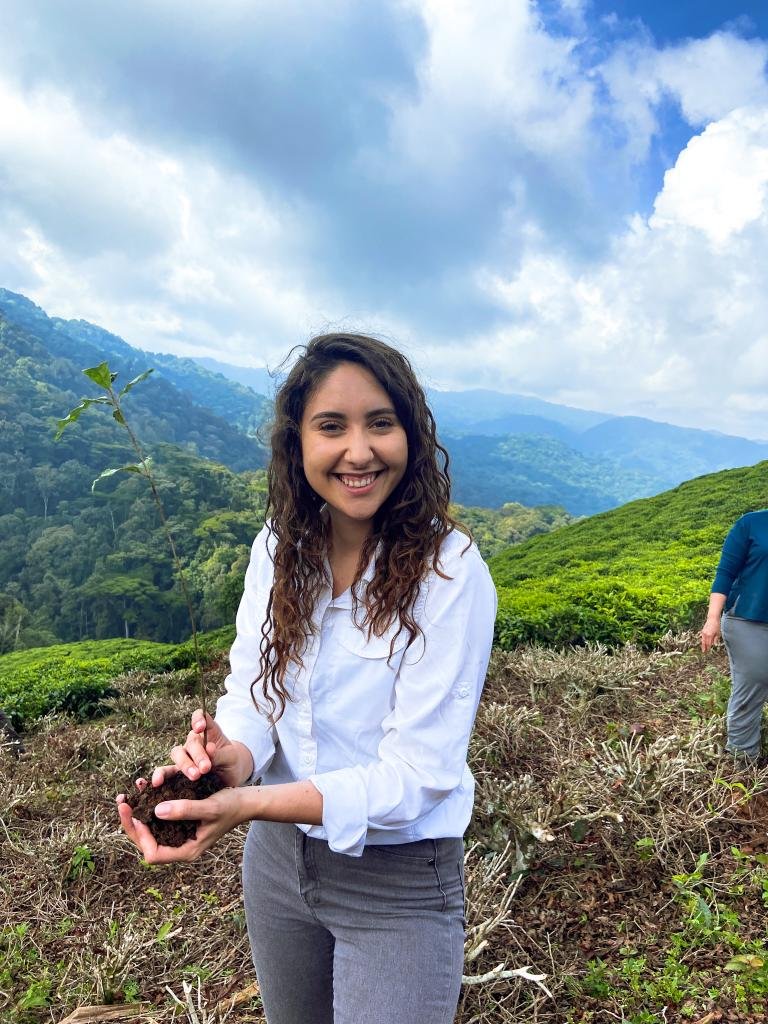 A young woman with curly hair smiling at the camera while holding soil and planting a small tree in a lush green mountainous landscape under a partly cloudy sky.