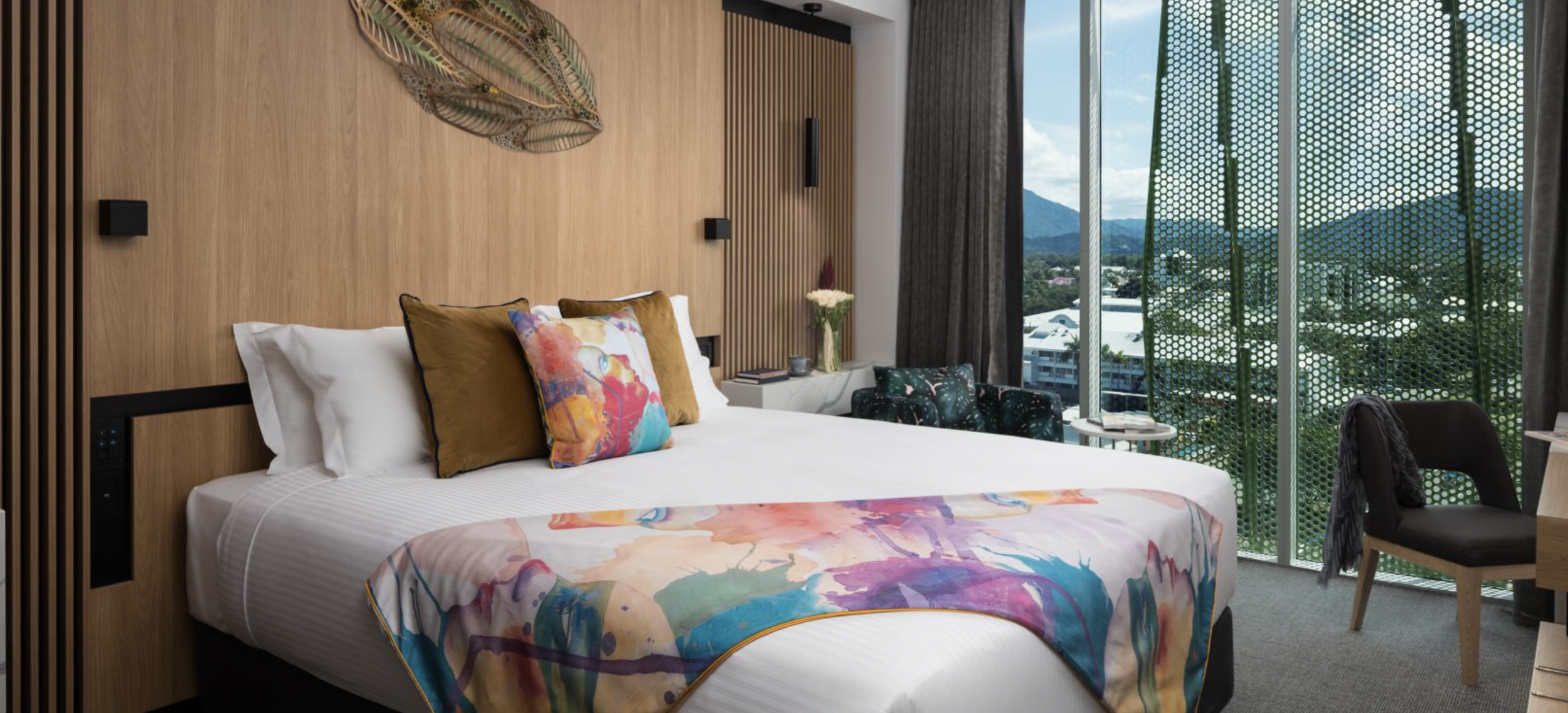 Modern hotel room with a bed fitted with white linens, colorful abstract throw blanket, and decorative pillows, against a wooden headboard with a scenic view of mountains through large windows covered partly by decorative green panels.