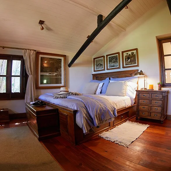 Cozy bedroom with wooden bed frame, blue and white bedding, nightstand with lamp, framed pictures on the wall, window with curtains, and a sloped ceiling with a black beam.